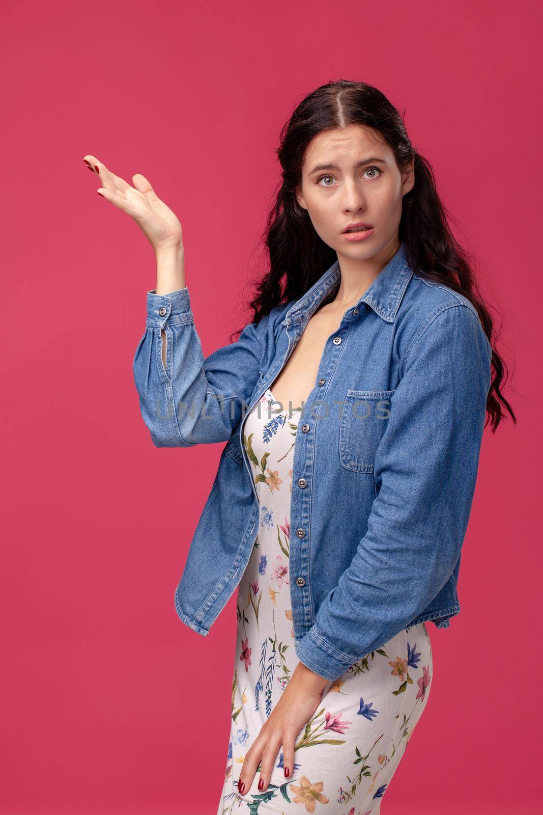 Portrait of a pretty young female in a white dress with floral print and blue denim shirt standing on a pink wall background in studio. She is looking confused. People sincere emotions, lifestyle concept. Mockup copy space.