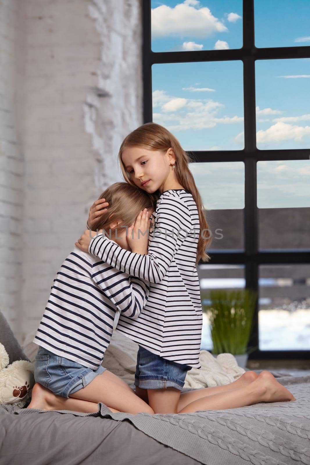 Two little sisters in striped t-shirts and denim shorts are sitting on the sofa in the bright spacious room. Little sister upset and crying. Oolder sister hugs younger and holds her head