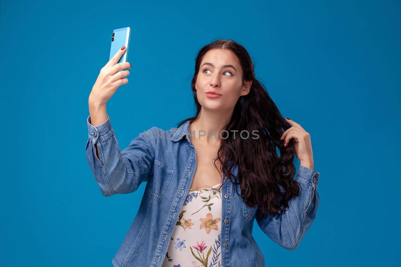 Young pretty female in a white dress with floral print and blue denim shirt is posing with a smartphone on a blue background. She is making a selfie. People sincere emotions, lifestyle concept. Mockup copy space.