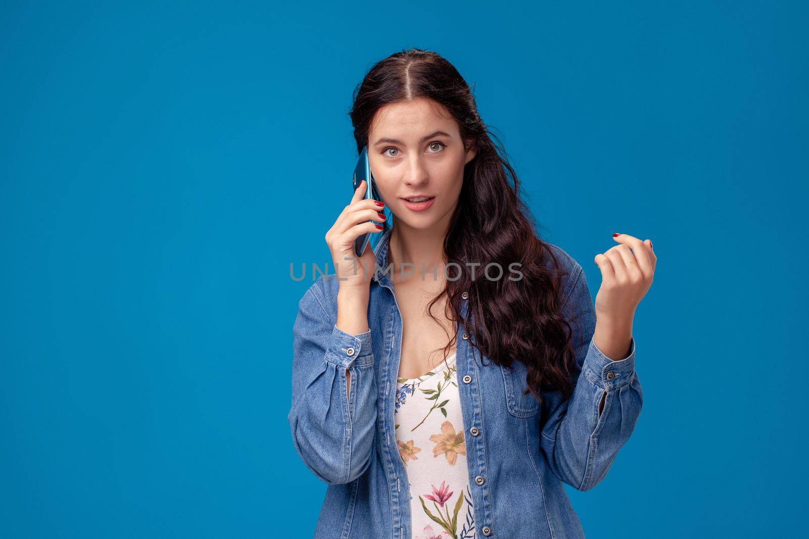 Young charming girl in a white dress with floral print and blue denim shirt is posing with a smartphone on a blue background. She is talking to someone. People sincere emotions, lifestyle concept. Mockup copy space.