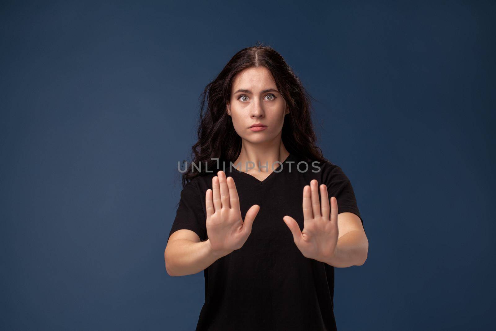 Portrait of disappointed female with curly hair dressed in a black t-shirt. She act like she is asking someone to stop, standing on a gray background.