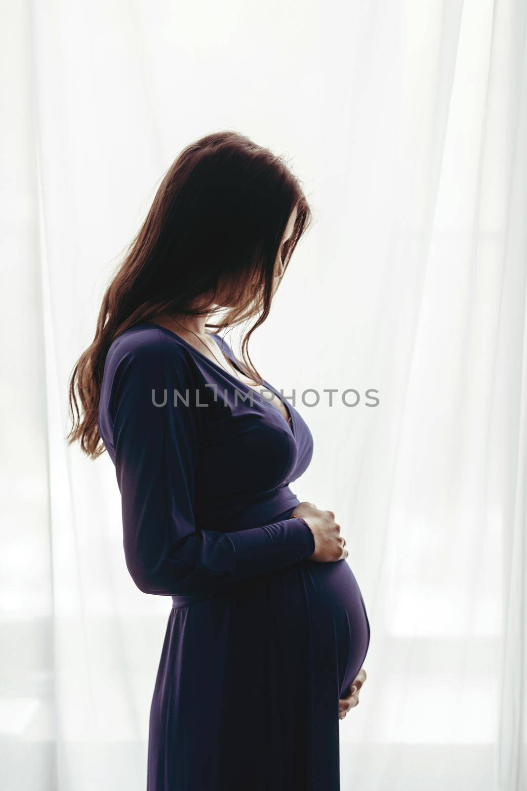 Pregnant woman in a blue dress at home standing against the window. Silhouette of a woman