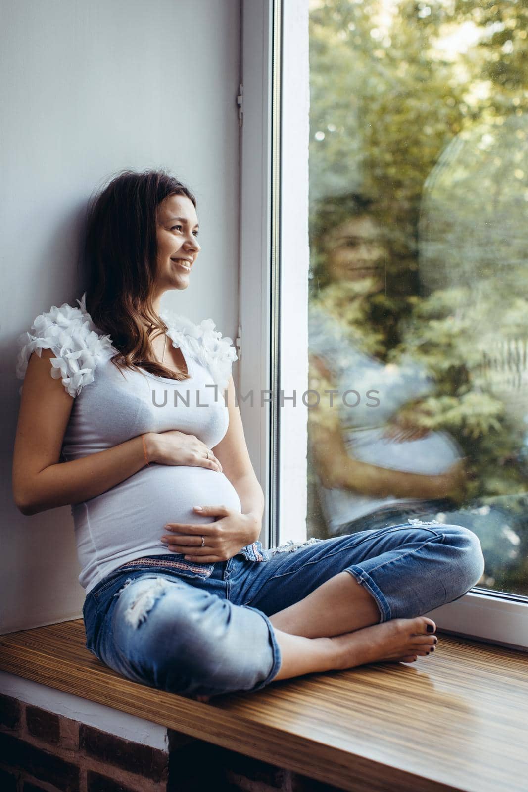 Beautiful pregnant brunette girl sitting on the windowsill, holding his stomach. Outside, the greens, the girl white T-shirt and ripped jeans Looking out the window, smiling.
