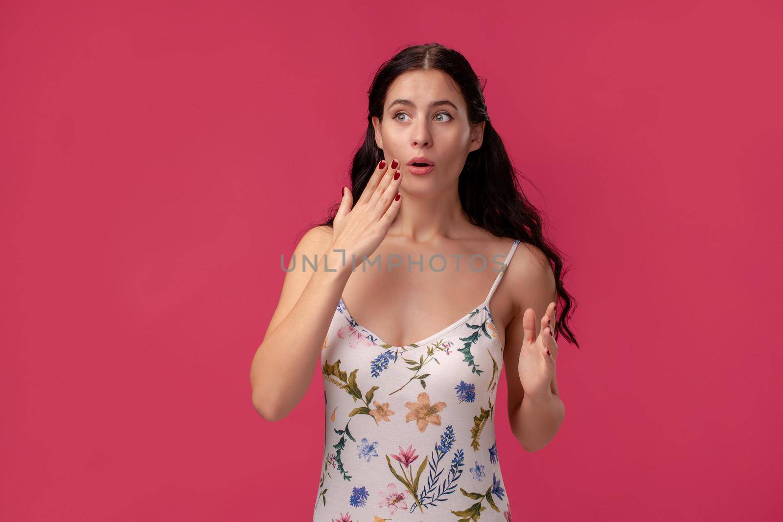 Portrait of a beautiful girl in a white dress with floral print standing on a pink wall background in studio. She is wondered and looking at someone. People sincere emotions, lifestyle concept. Mockup copy space.
