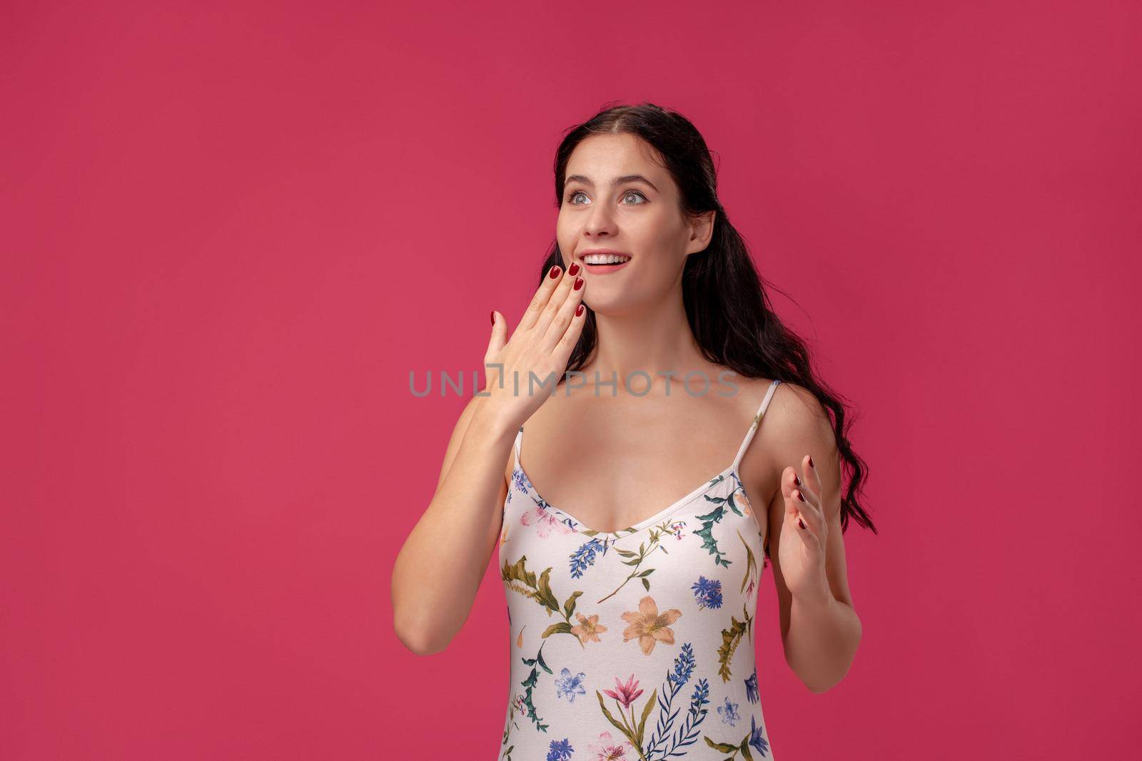 Portrait of a good-looking girl in a white dress with floral print standing on a pink wall background in studio. She is shocked, covering her mouth with her hand and smiling. People sincere emotions, lifestyle concept. Mockup copy space.