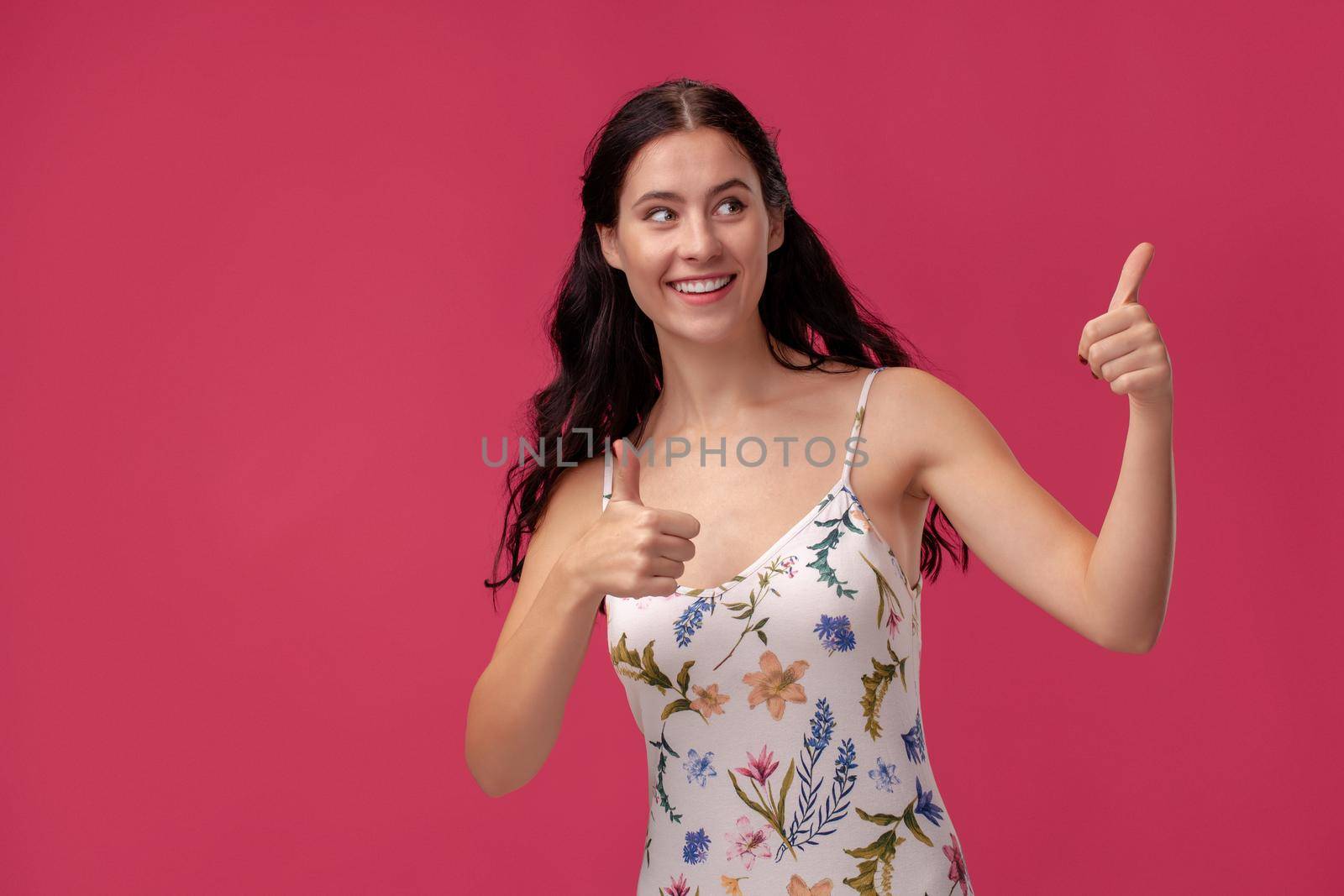 Portrait of a brunette young woman in a white dress with floral print standing on a pink wall background in studio. She showing ok and smiling to someone. People sincere emotions, lifestyle concept. Mockup copy space.