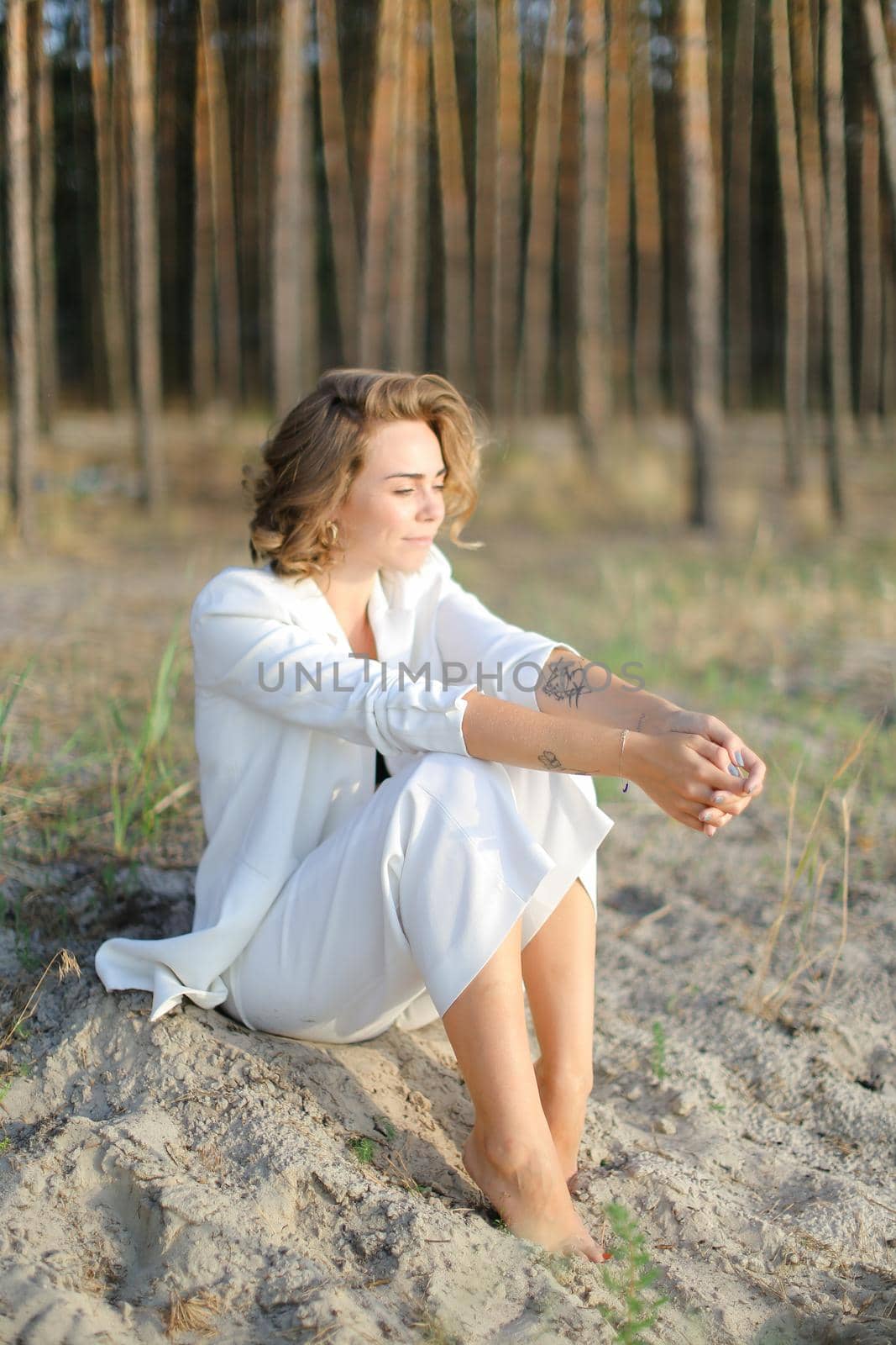 Young blonde girl sitting on sand beach and wearing white clothes.