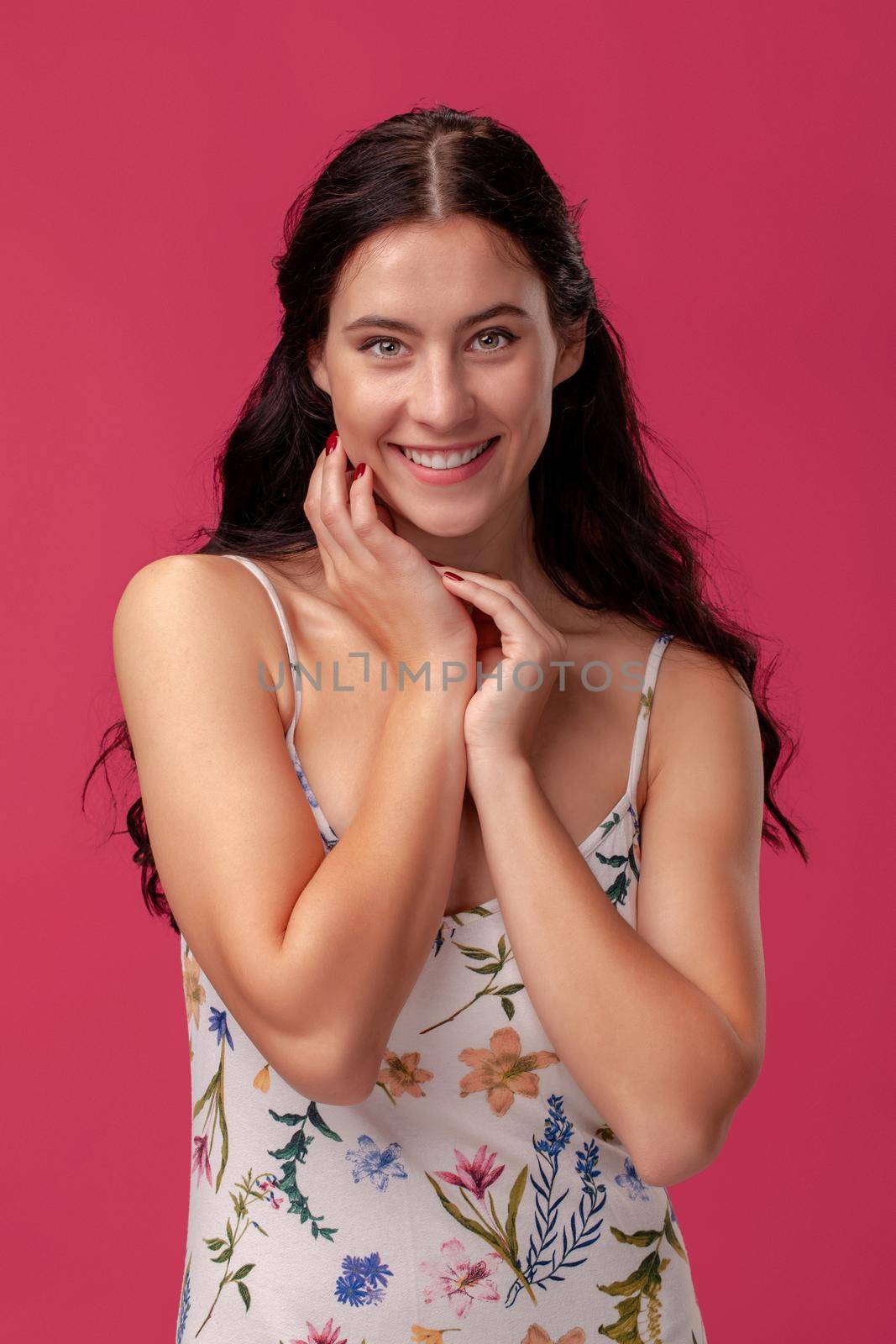 Portrait of a smiling brunette woman with a long curly hair, wearing in a white dress with floral print and standing on a pink wall background in studio. She folded her hands on a chest and looking at the camera. People sincere emotions, lifestyle concept. Mockup copy space.