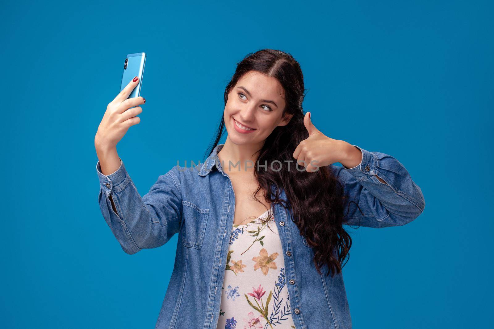 Young pretty girl in a white dress with floral print and blue denim shirt is posing with a smartphone on a blue background. She is making a selfie. People sincere emotions, lifestyle concept. Mockup copy space.