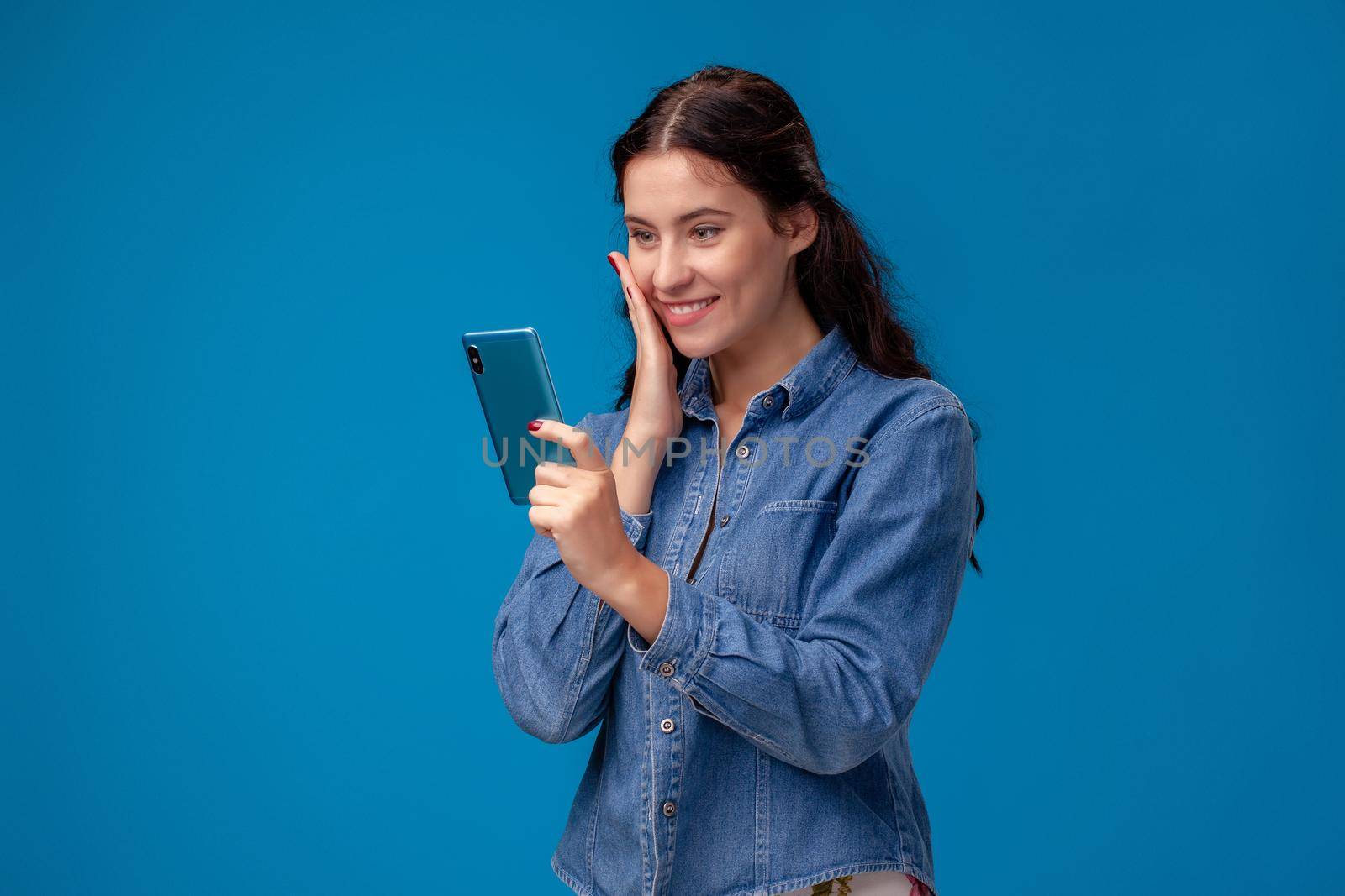 Young brunette lady in a blue denim shirt is posing with a smartphone on a blue background. She is chatting with someone and smiling. People sincere emotions, lifestyle concept. Mockup copy space.