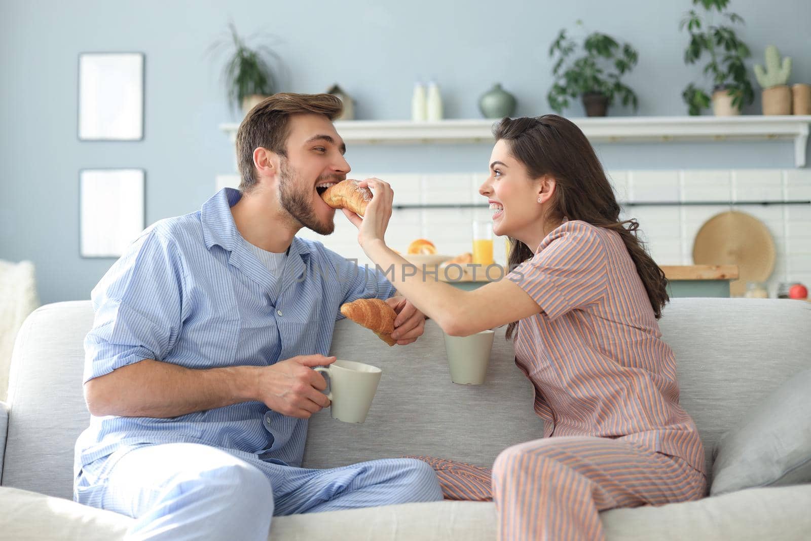 Happy young couple in pajamas in kitchen having breakfast, feeding each other a croissant. by tsyhun