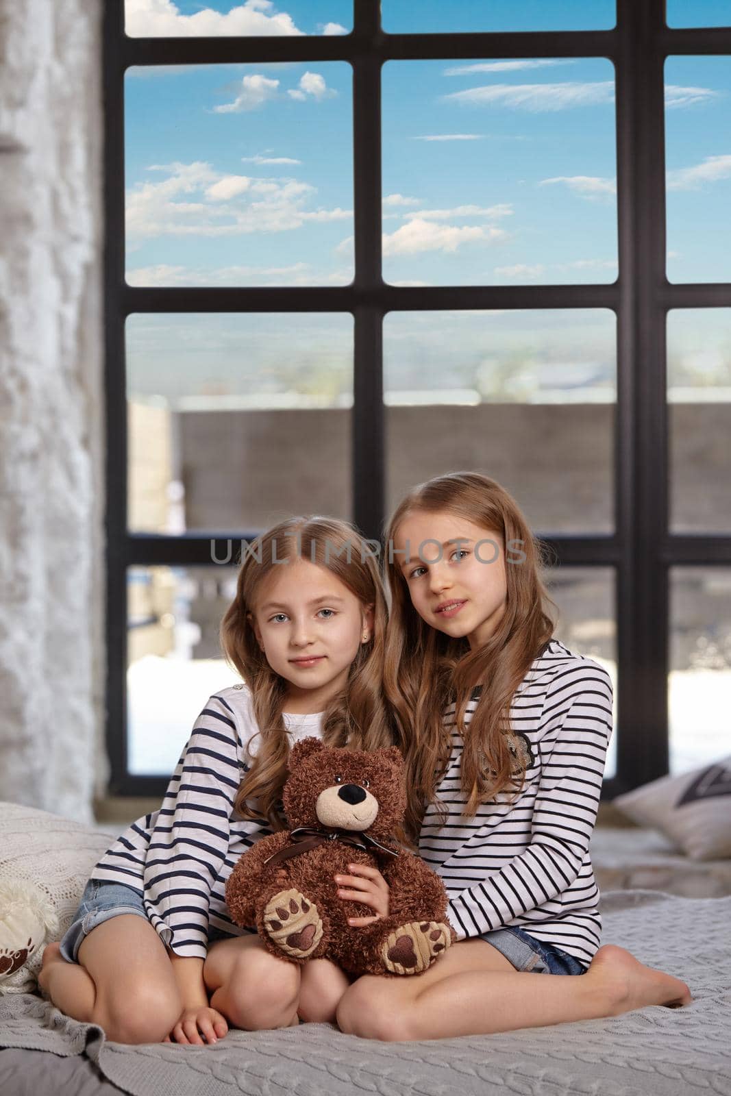 Cute little sisters in striped t-shirts are sitting on the sofa in the bright spacious room. They are holding little brown toy bear between them