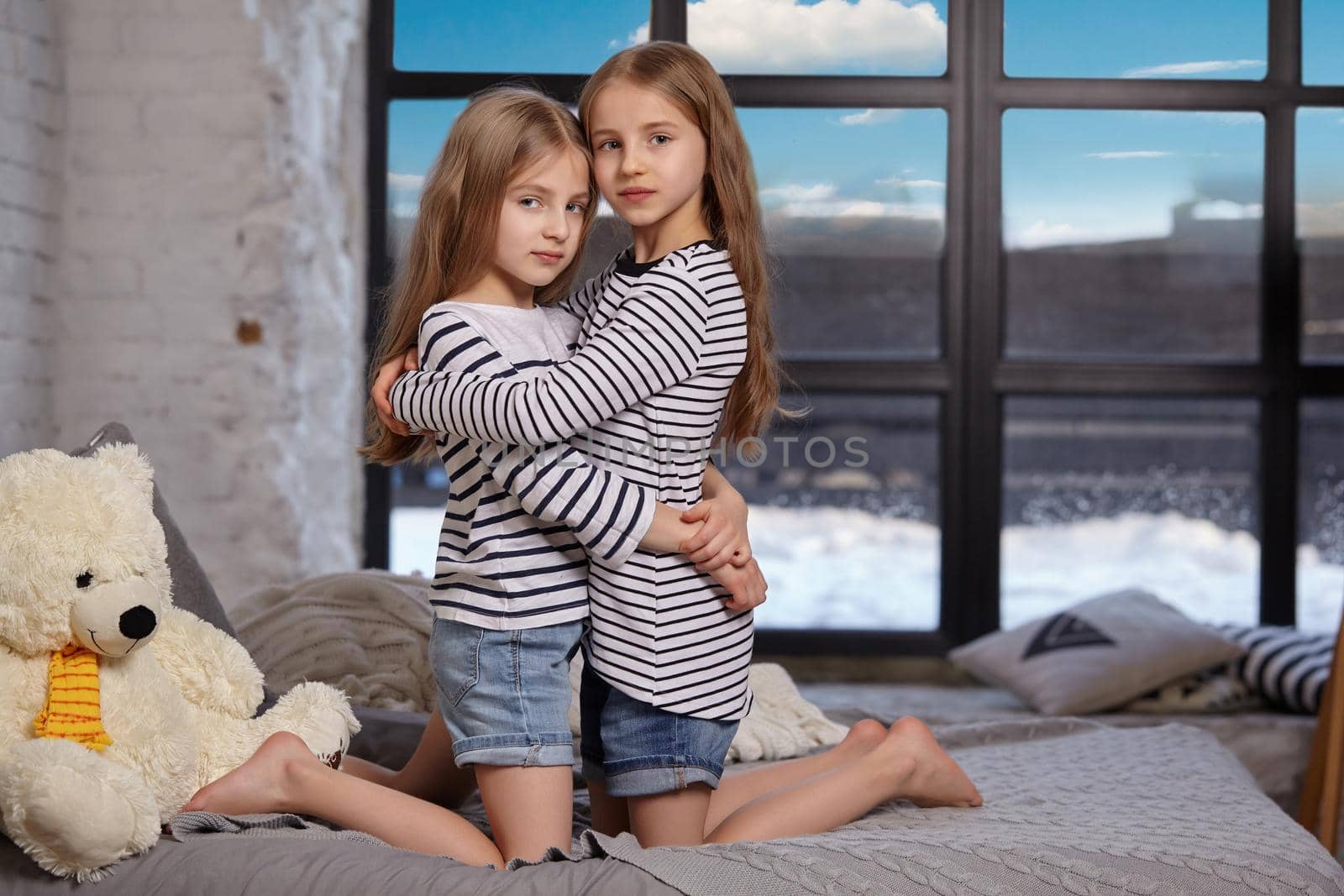 Close up photo of two little sisters in striped t-shirts are sitting on the sofa in the bright spacious room. They hug each other