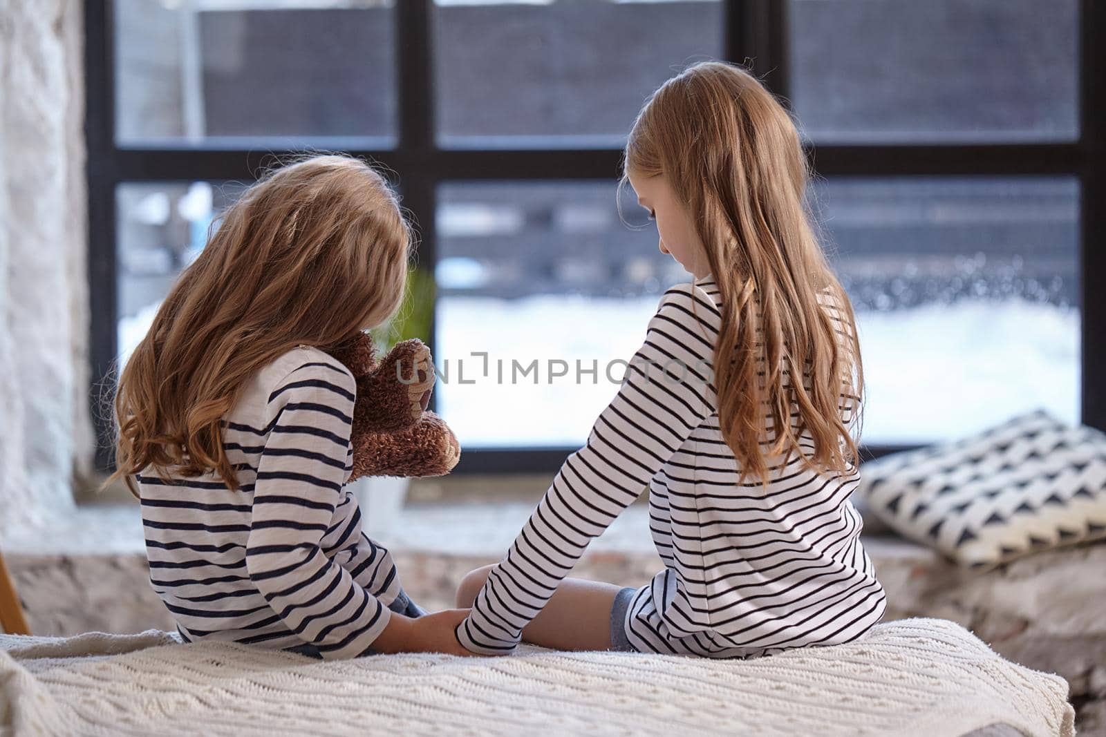 Two cute little sisters sitting on the bed in their bedroom before big window back to camera.