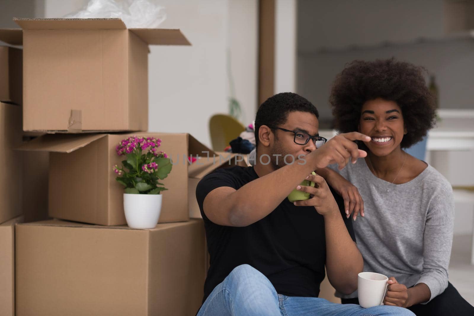 Relaxing in new house. Cheerful young African American couple sitting on the floor and drinking coffee while cardboard boxes laying all around them