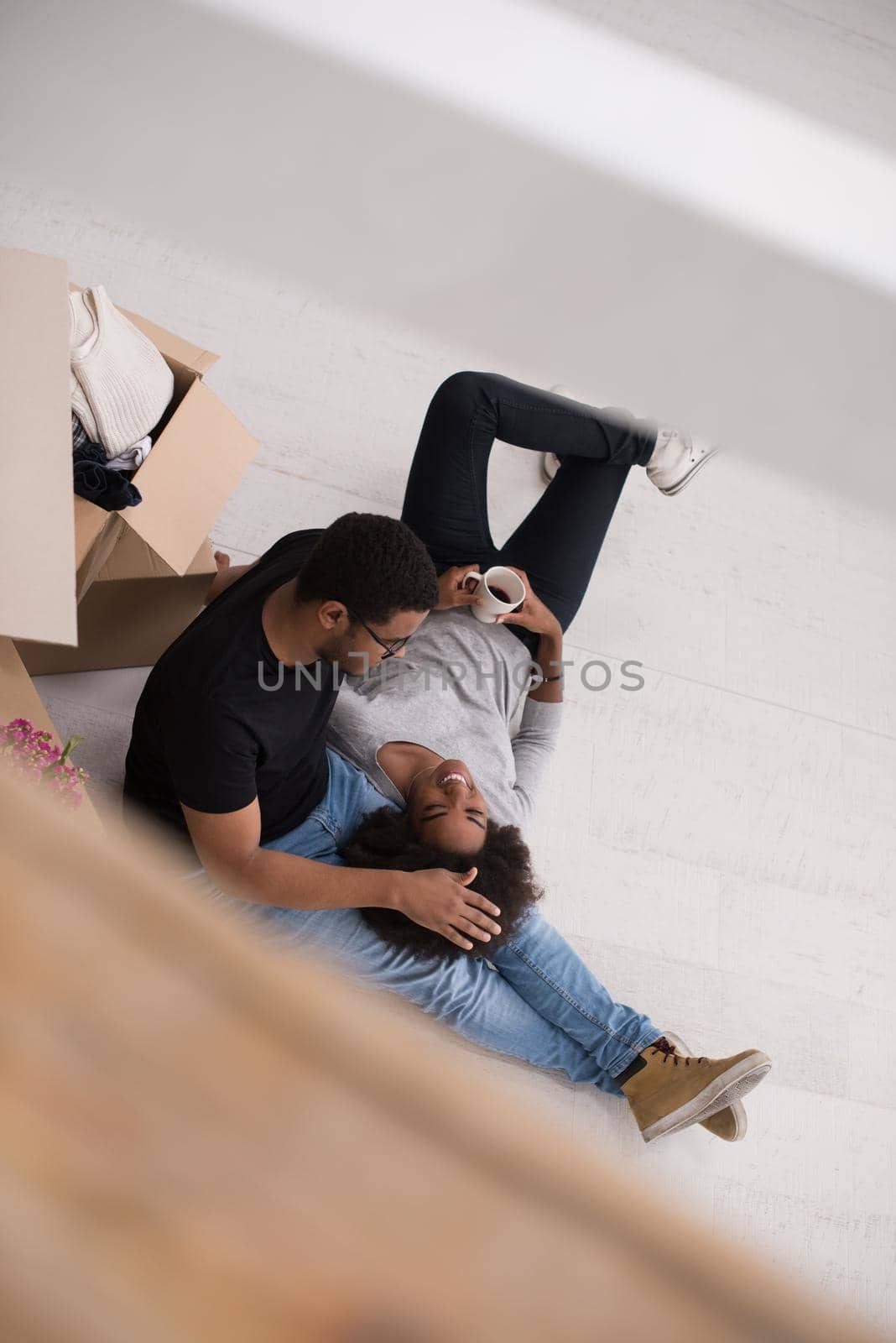 Relaxing in new house. Cheerful young African American couple sitting on the floor and drinking coffee while cardboard boxes laying all around them