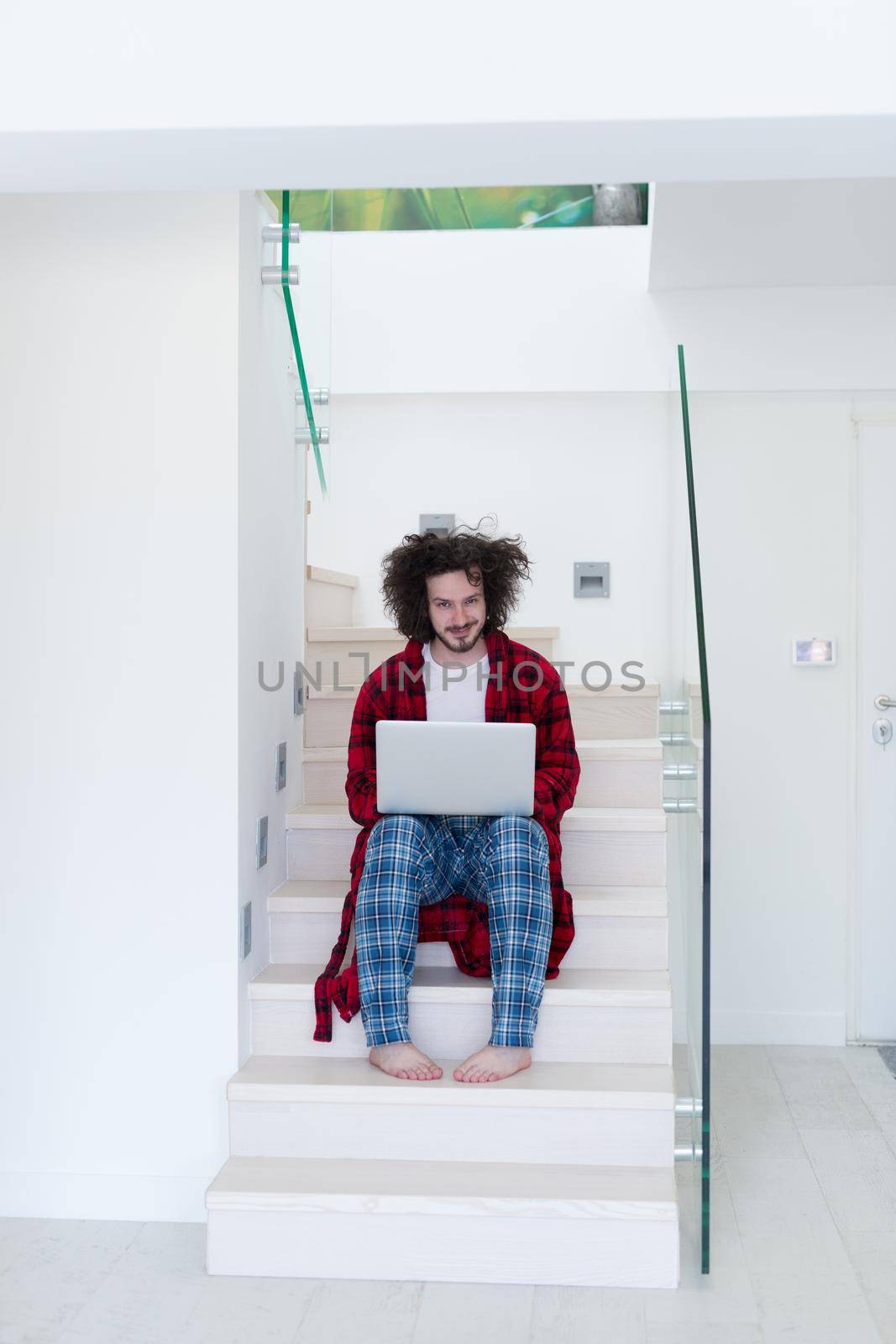 young freelancer in bathrobe working from home using laptop computer while sitting on stairs