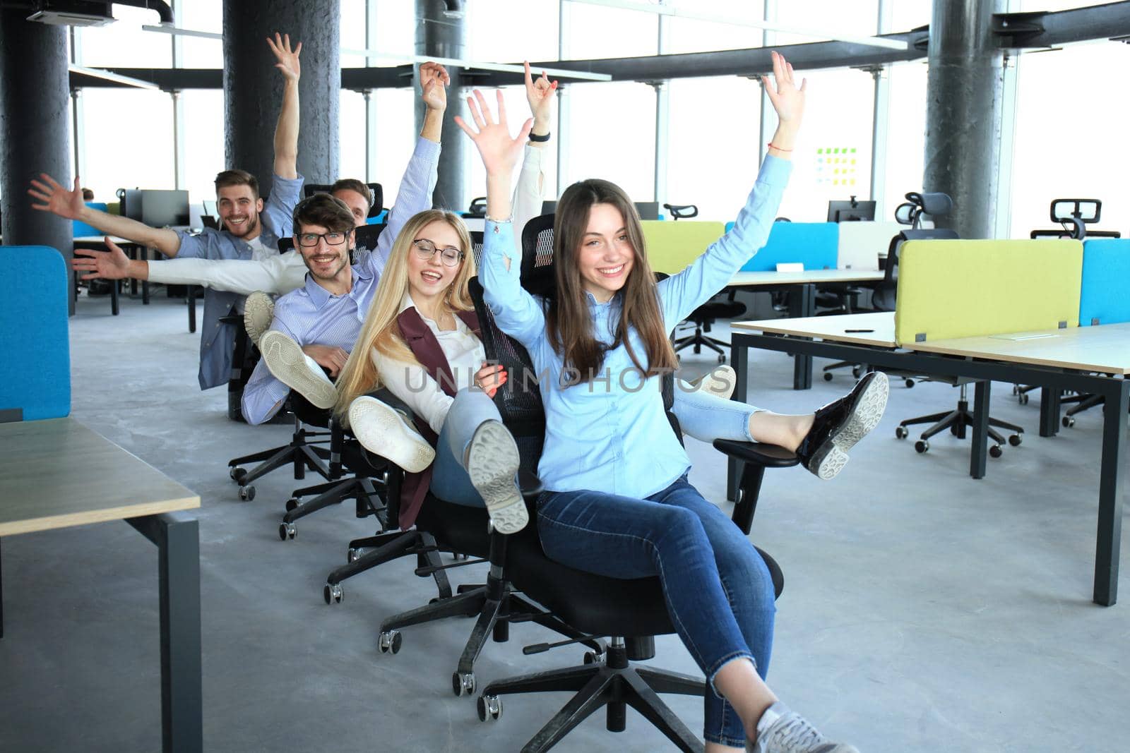 Young cheerful business people in smart casual wear having fun while racing on office chairs and smiling. by tsyhun