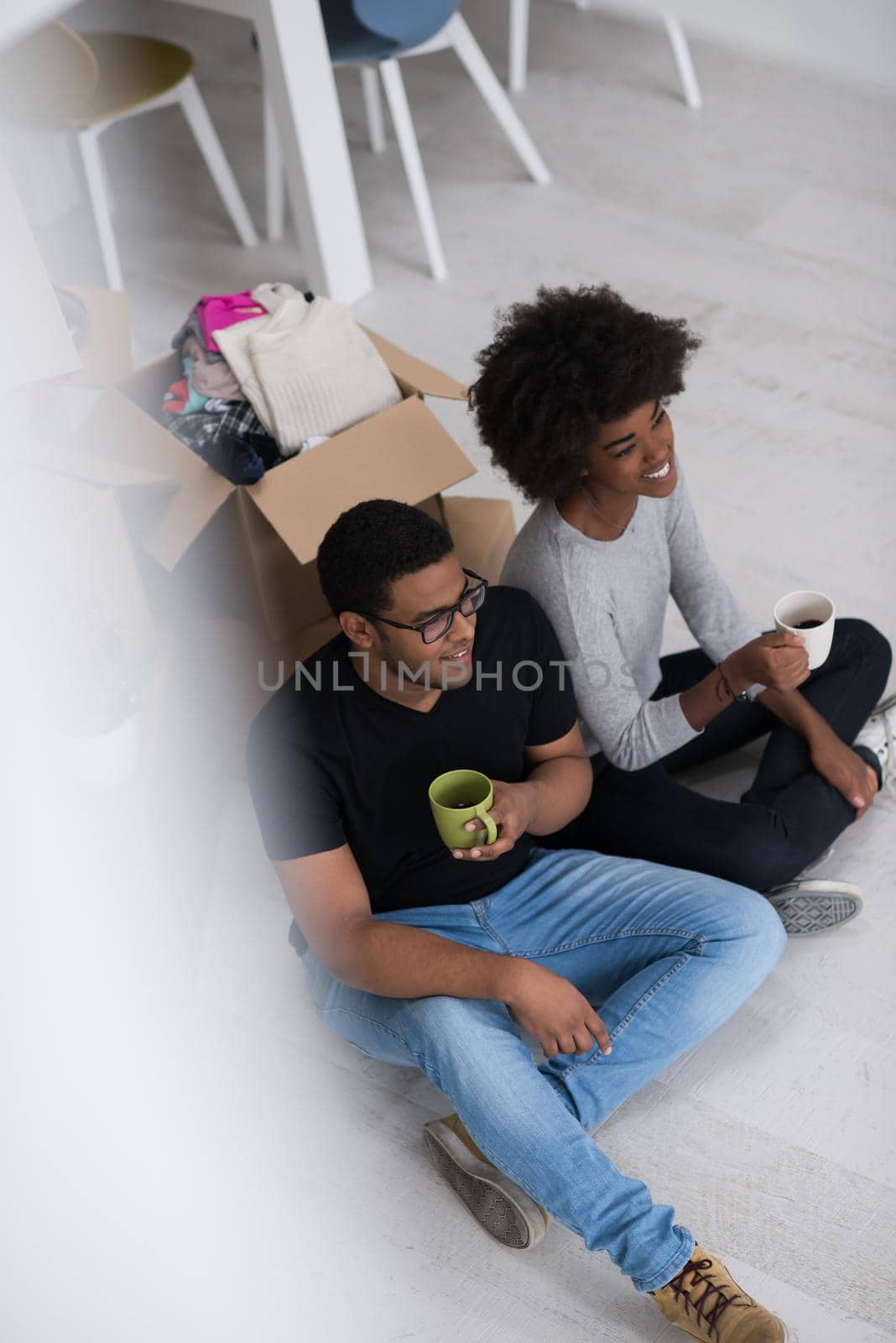 Relaxing in new house. Cheerful young African American couple sitting on the floor and drinking coffee while cardboard boxes laying all around them