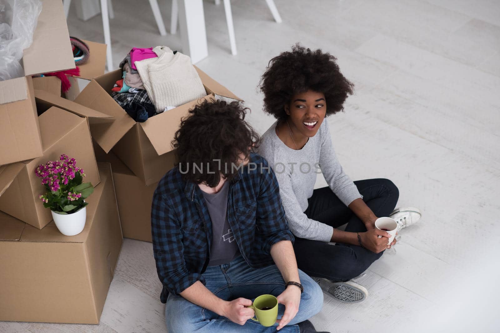 Relaxing in new house. Cheerful young multiethnic couple sitting on the floor and drinking coffee while cardboard boxes laying all around them