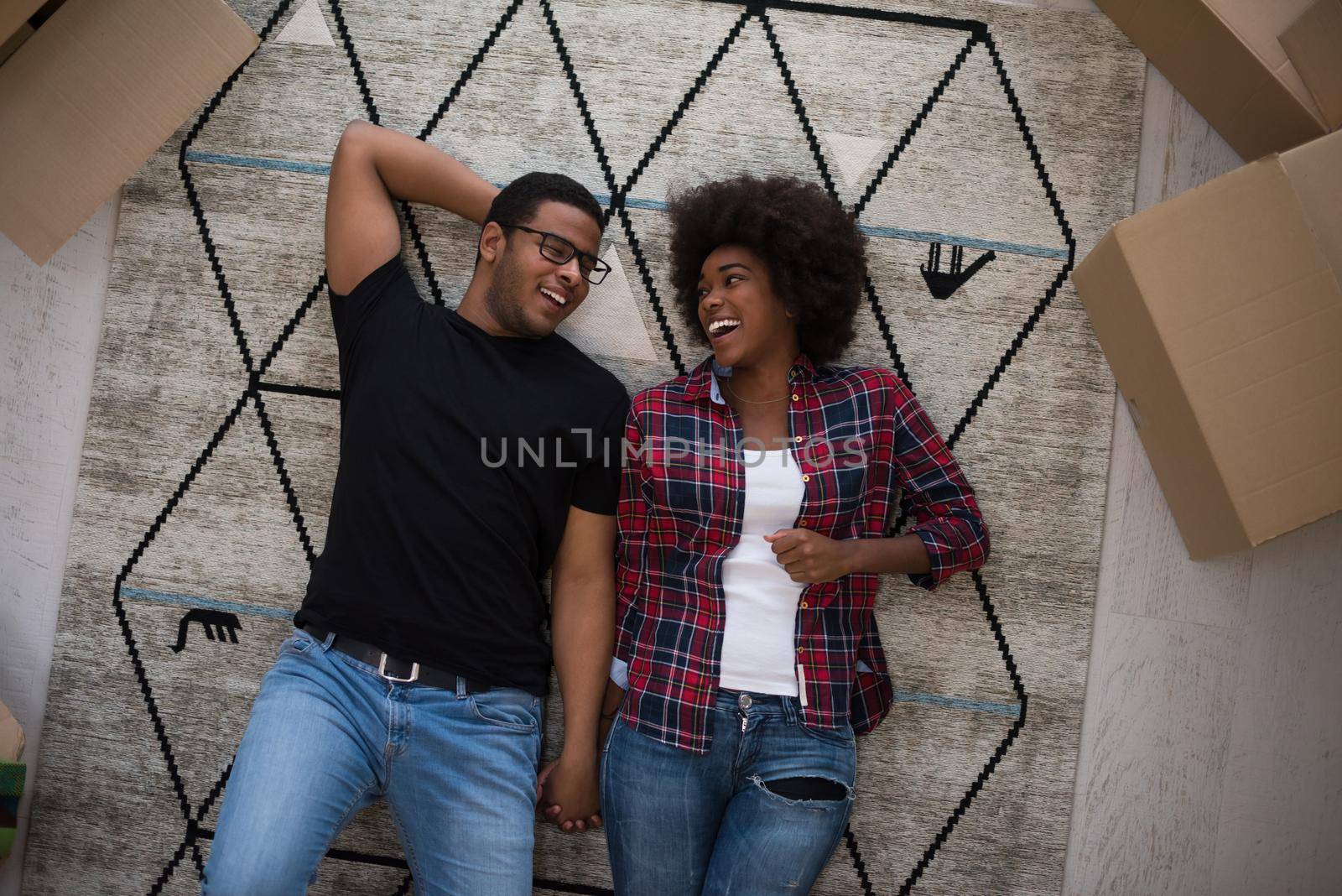 Top view of attractive young African American couple moving, holding hands, looking at camera and smiling while lying among cardboard boxes