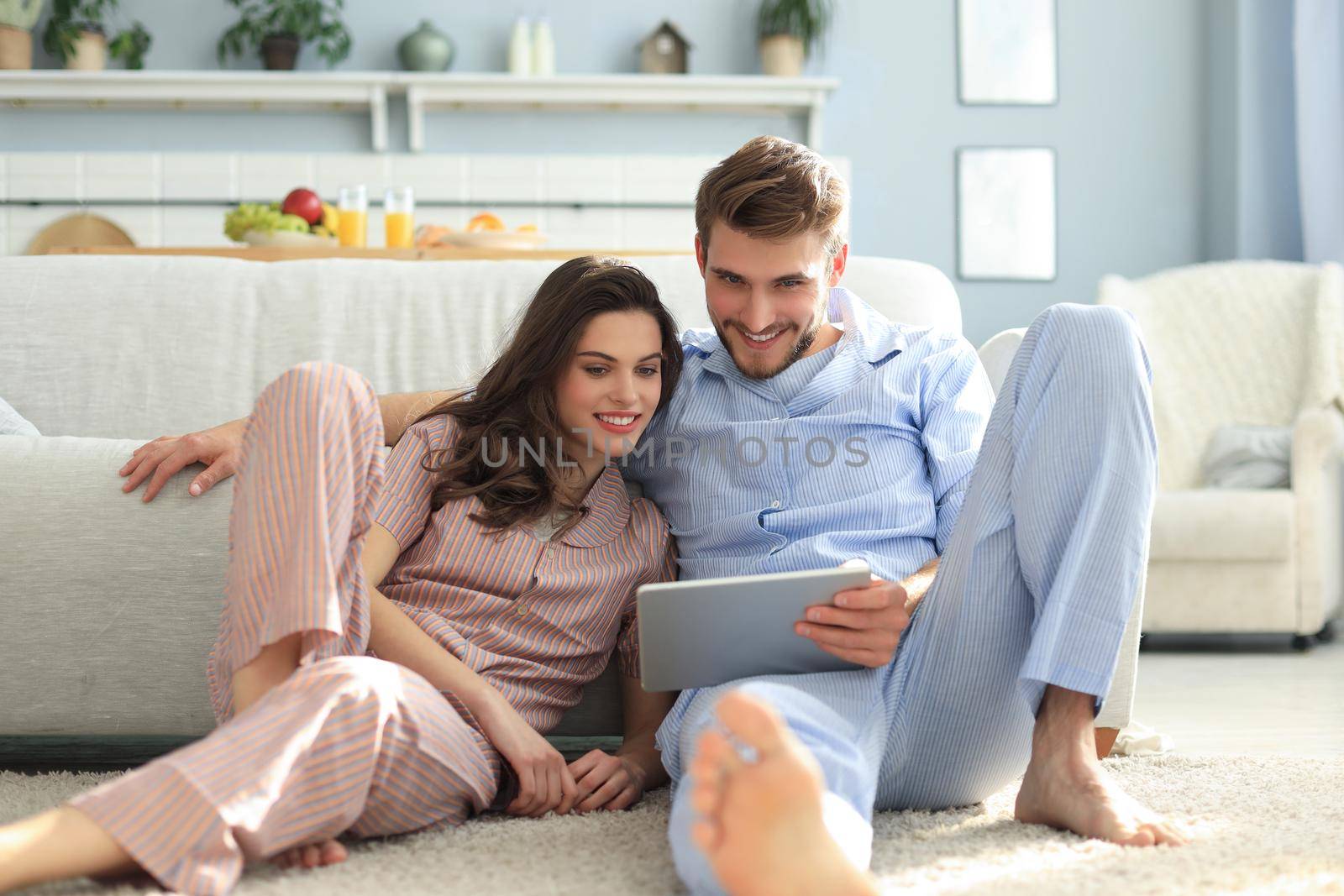 Young couple in pajamas watching media content online in a tablet sitting on the floor in the living room