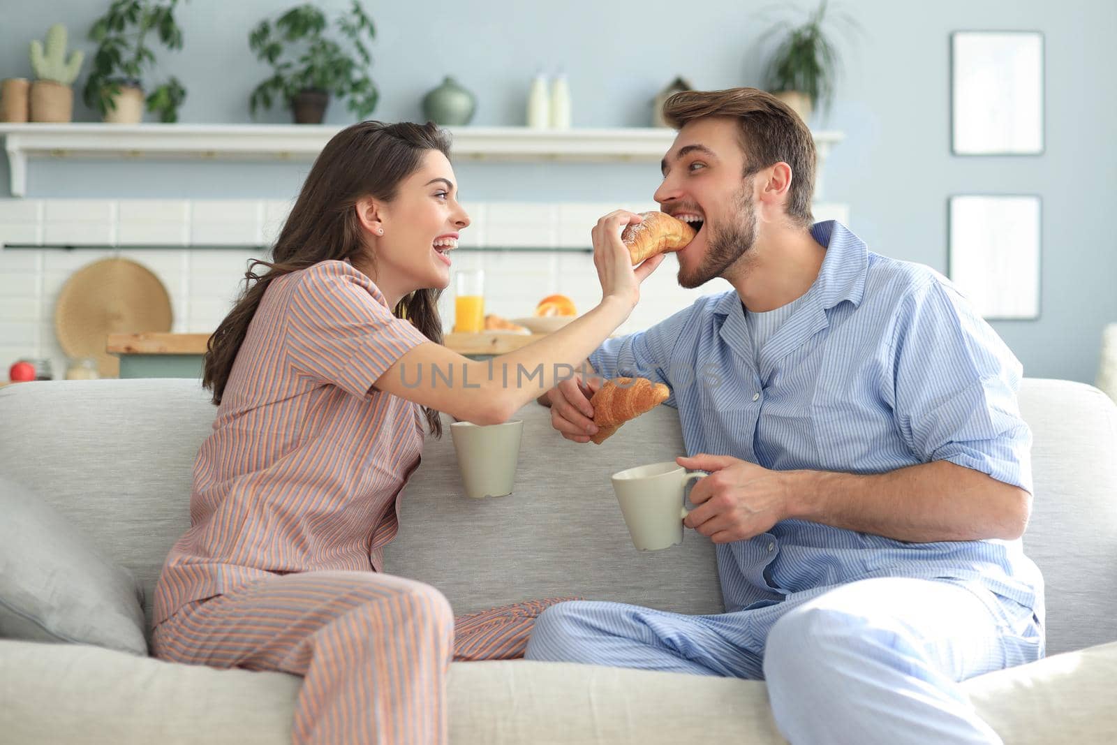 Happy young couple in pajamas in kitchen having breakfast, feeding each other a croissant. by tsyhun