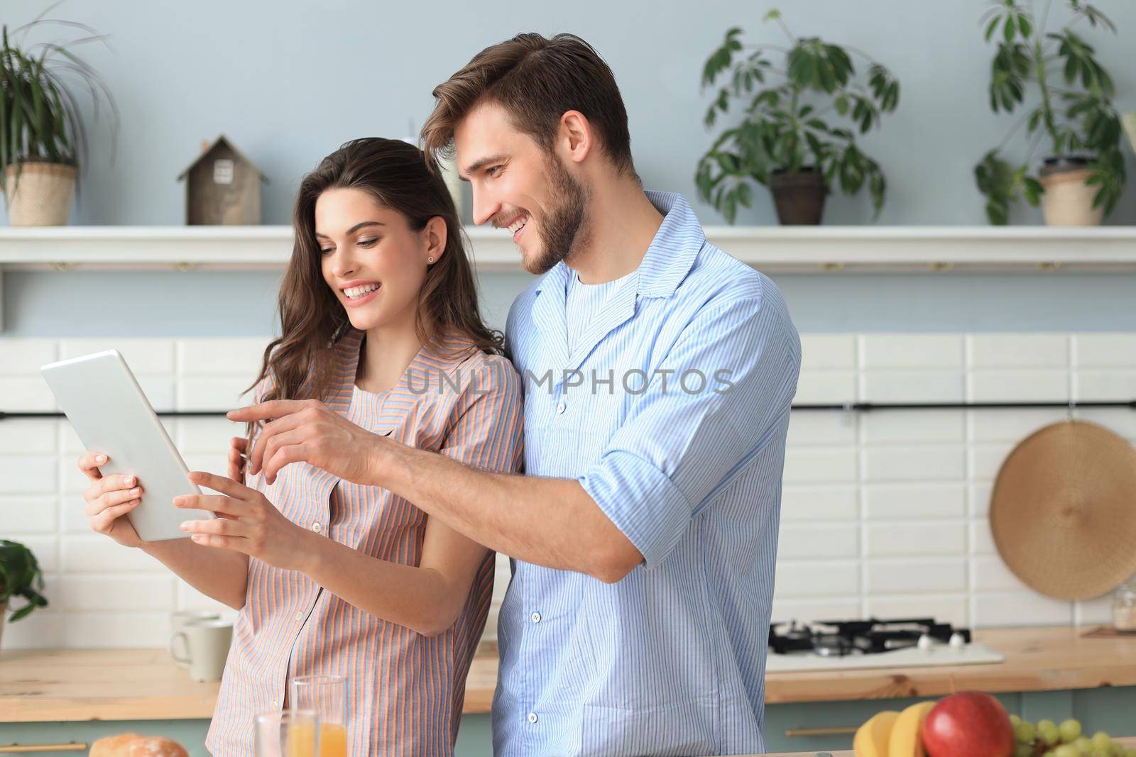 Happy young couple in pajamas watching online content in a didital tablet and smiling in kitchen at home