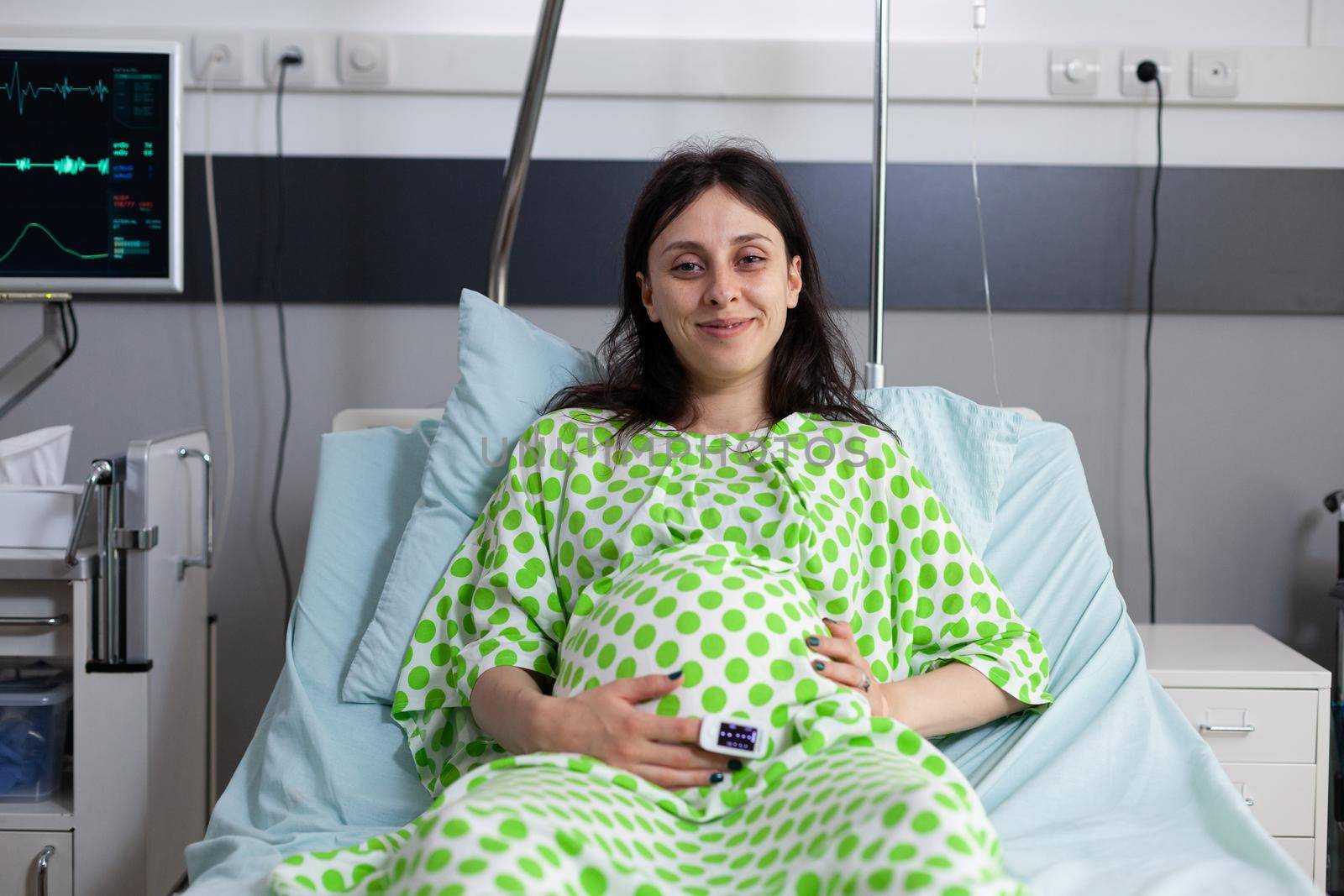 Portrait of pregnant woman sitting in hospital ward bed, looking at camera. Young person smiling, waiting for child delivery and parenthood at medical clinic. Adult with baby bump