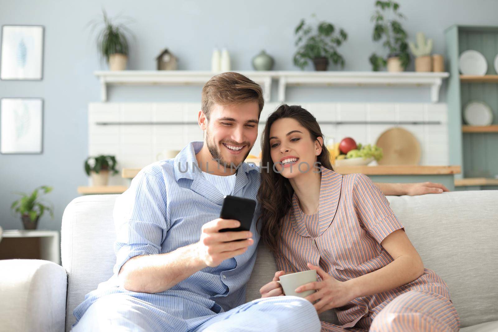 Young couple in pajamas watching online content in a smart phone sitting on a sofa at home in the living room. by tsyhun