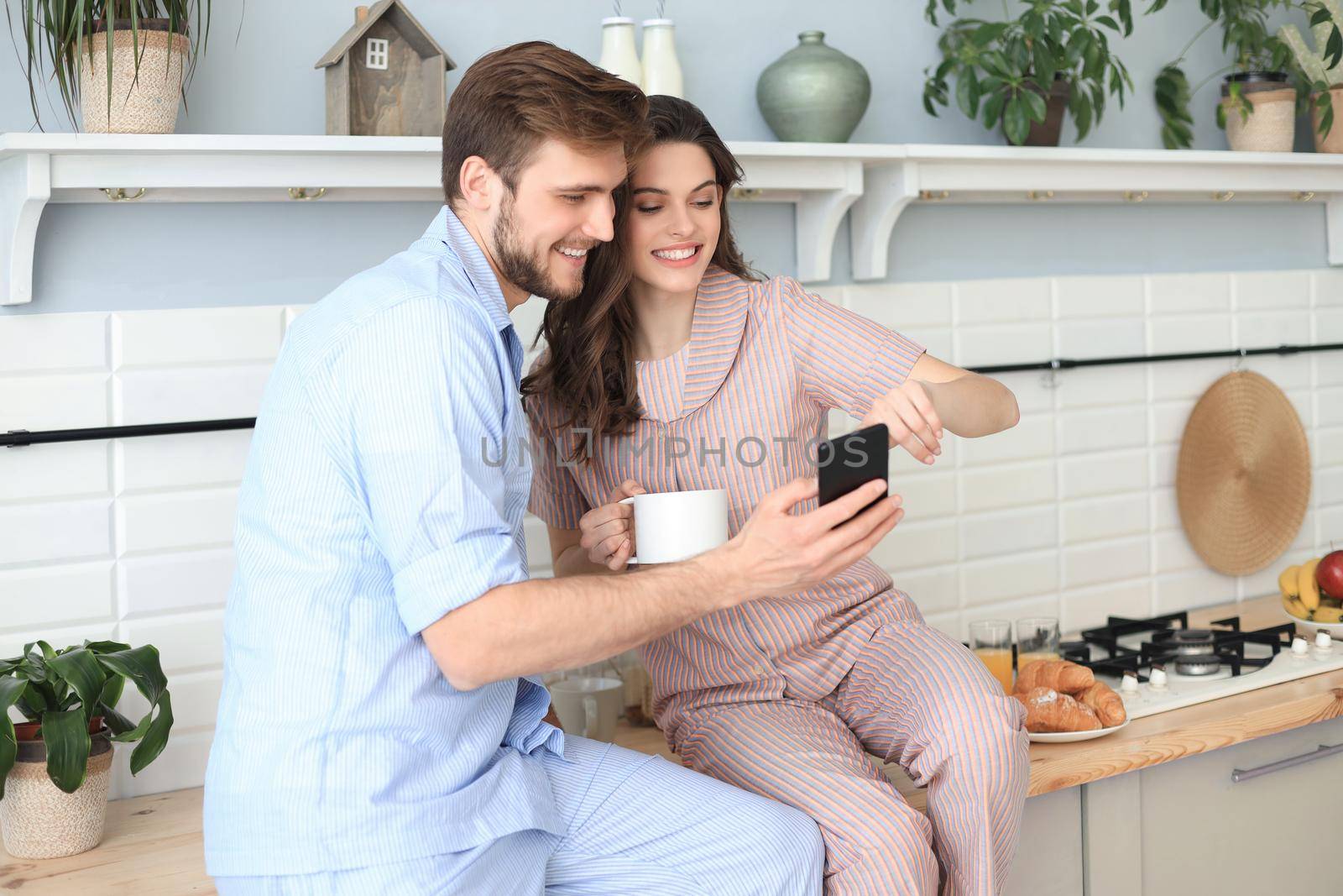 Happy young couple in pajamas watching online content in a smart phone and smiling while cooking in kitchen at home