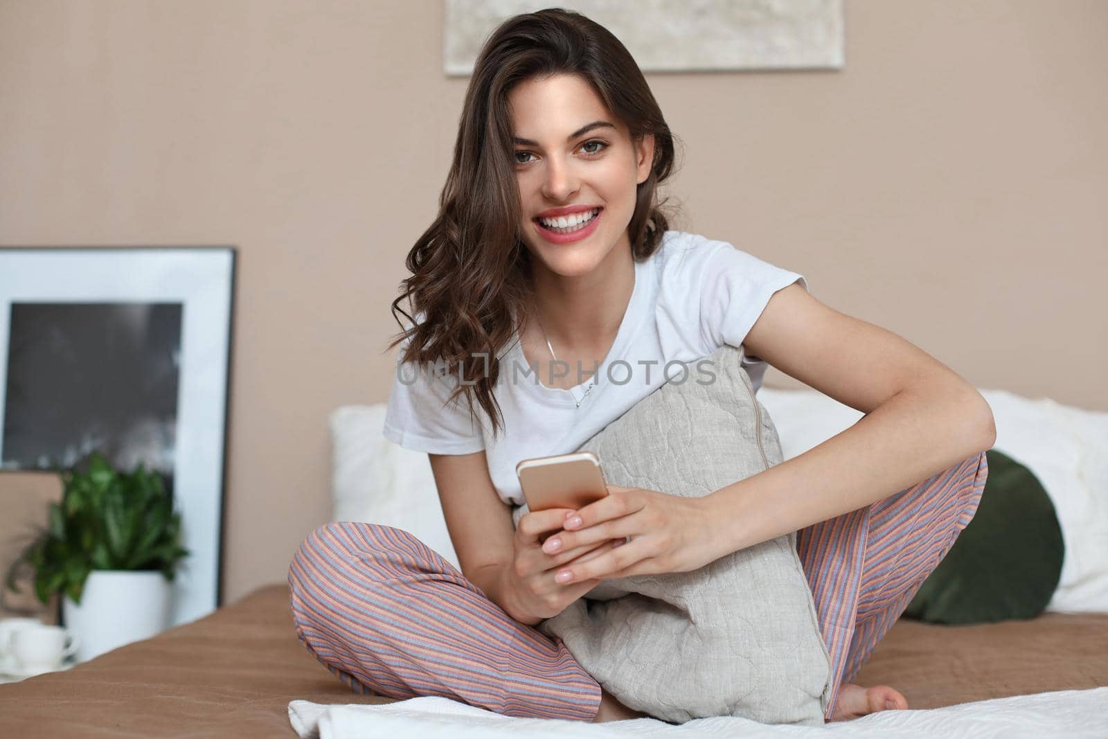 Beautiful young smiling brunette woman sitting in bed and using a phone in her bedroom