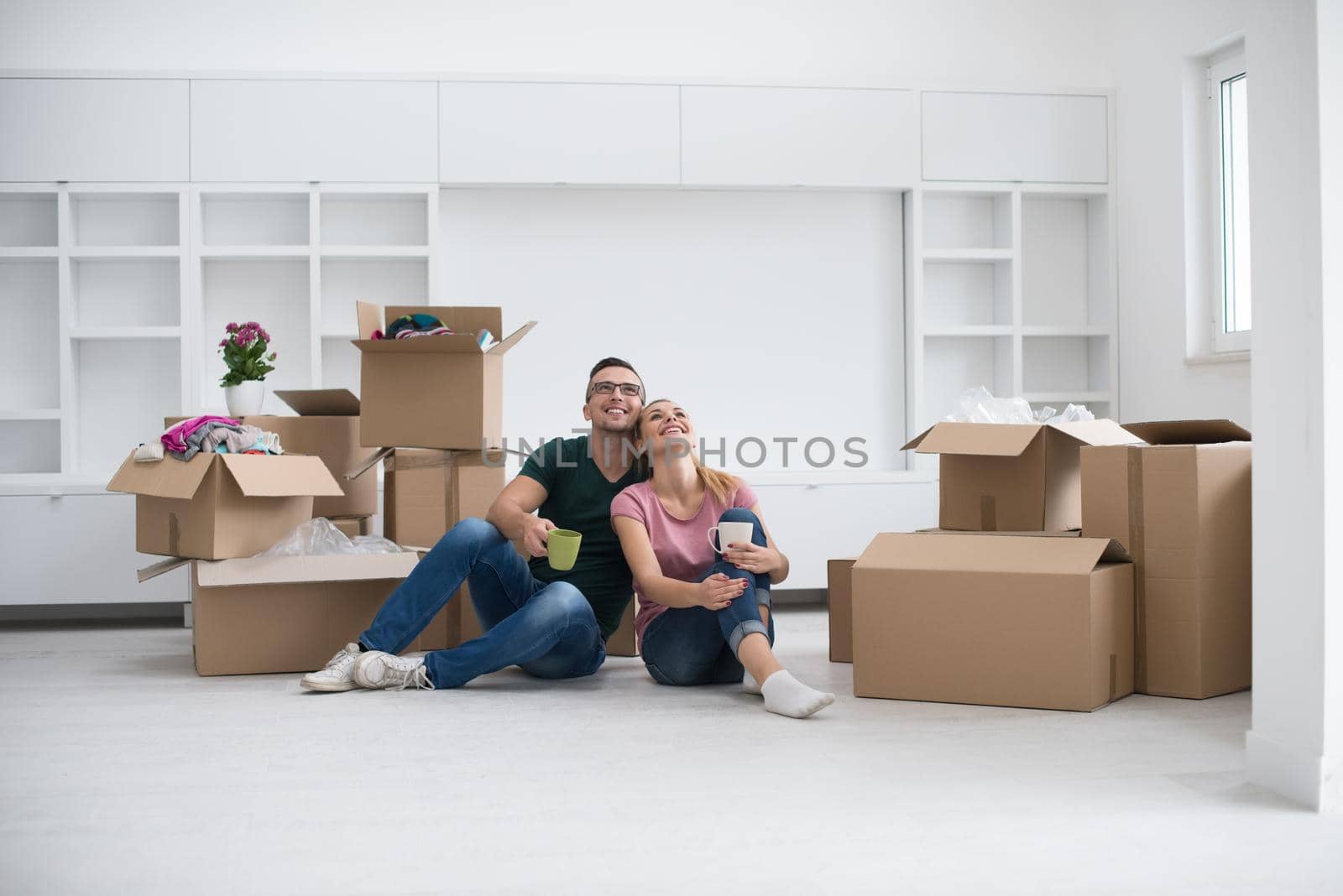 Relaxing in new house. Cheerful young couple sitting on the floor and drinking coffee while cardboard boxes laying all around them