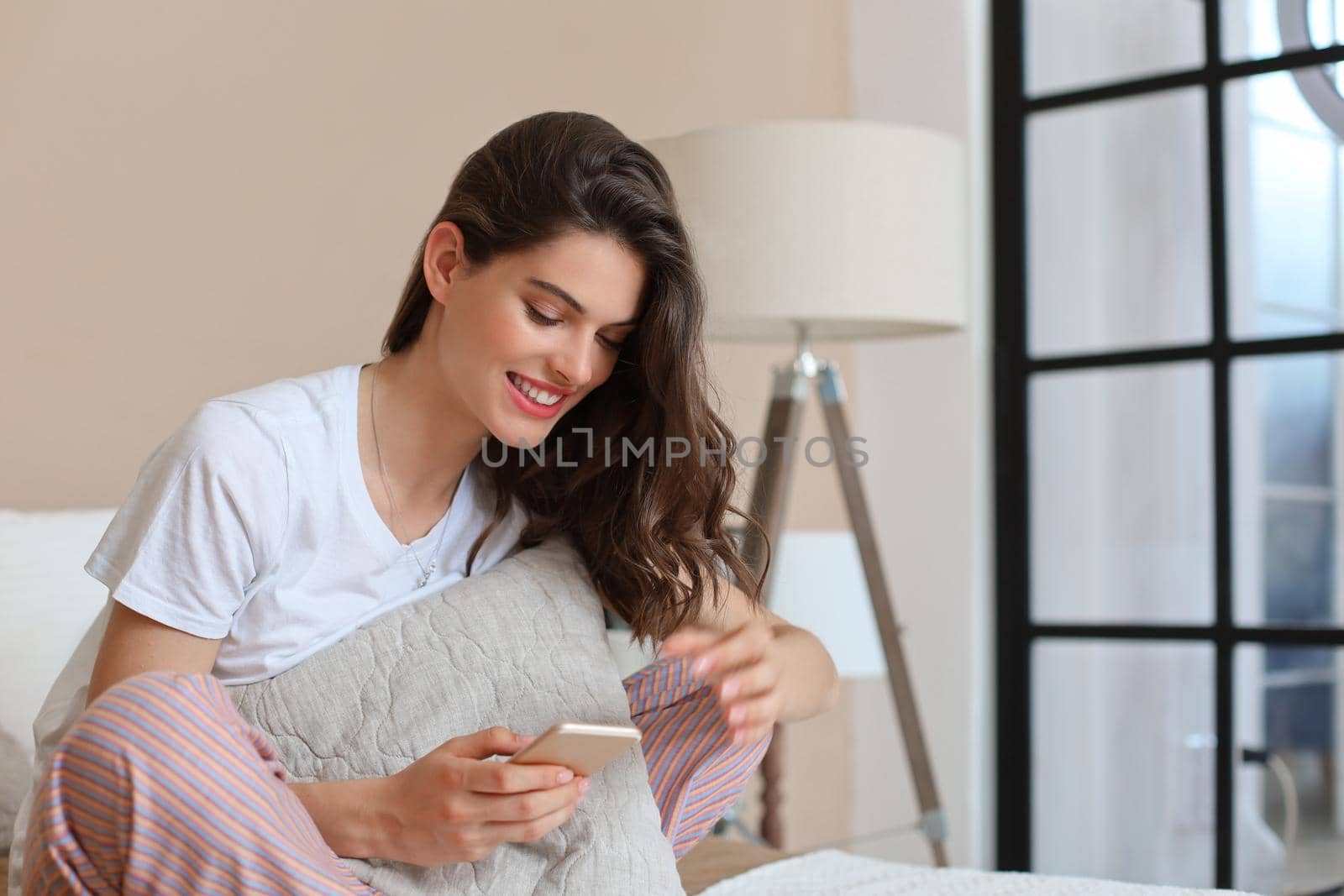 Beautiful young smiling brunette woman sitting in bed and using a phone in her bedroom