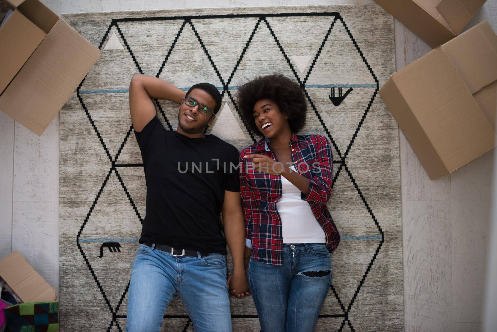 Top view of attractive young African American couple moving, holding hands, looking at camera and smiling while lying among cardboard boxes