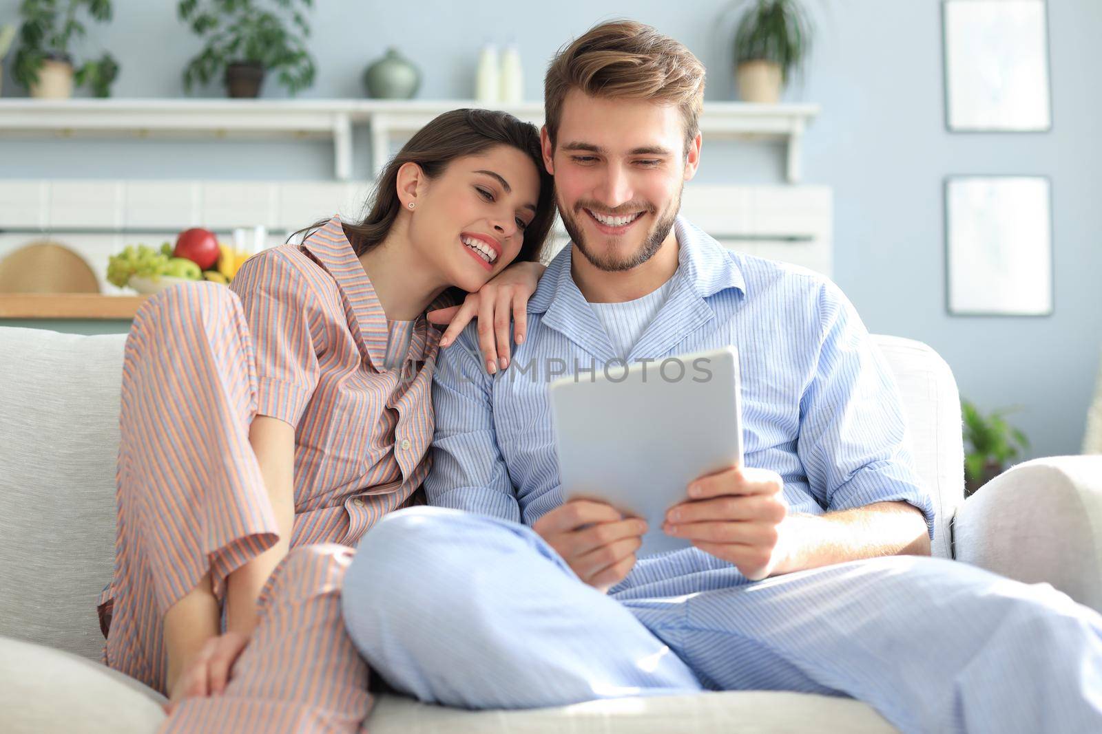 Young couple in pajamas watching media content online in a tablet sitting on a sofa in the living room. by tsyhun