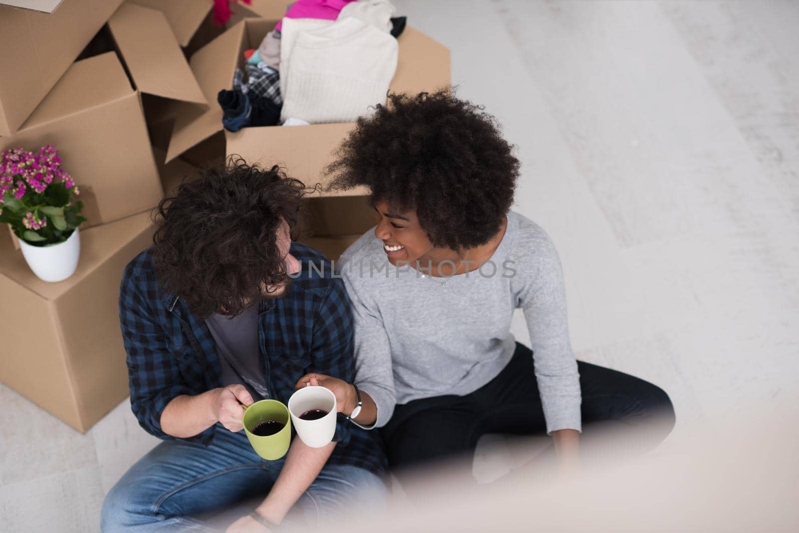 Relaxing in new house. Cheerful young multiethnic couple sitting on the floor and drinking coffee while cardboard boxes laying all around them