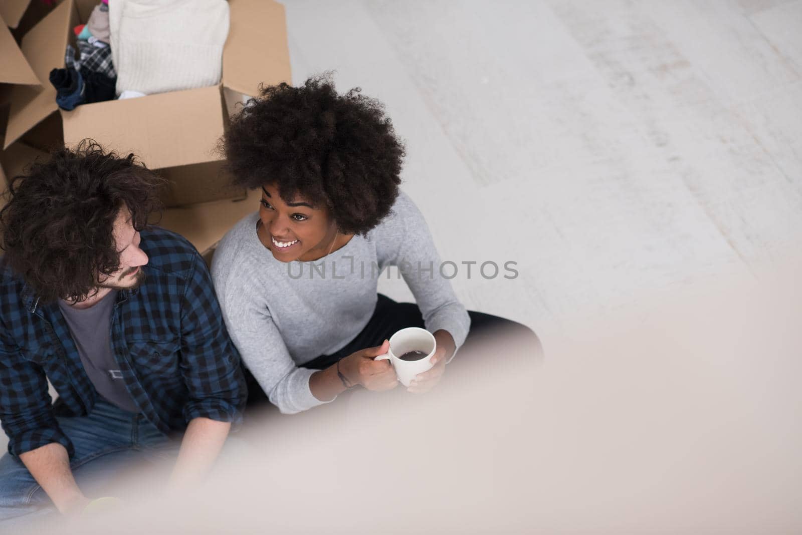 Relaxing in new house. Cheerful young multiethnic couple sitting on the floor and drinking coffee while cardboard boxes laying all around them