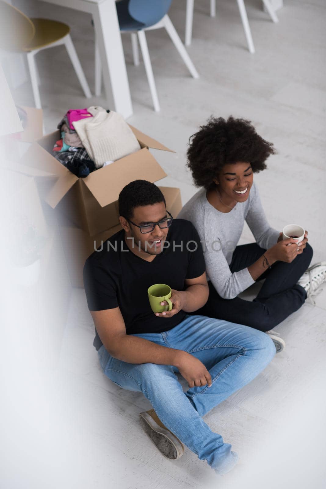 Relaxing in new house. Cheerful young African American couple sitting on the floor and drinking coffee while cardboard boxes laying all around them