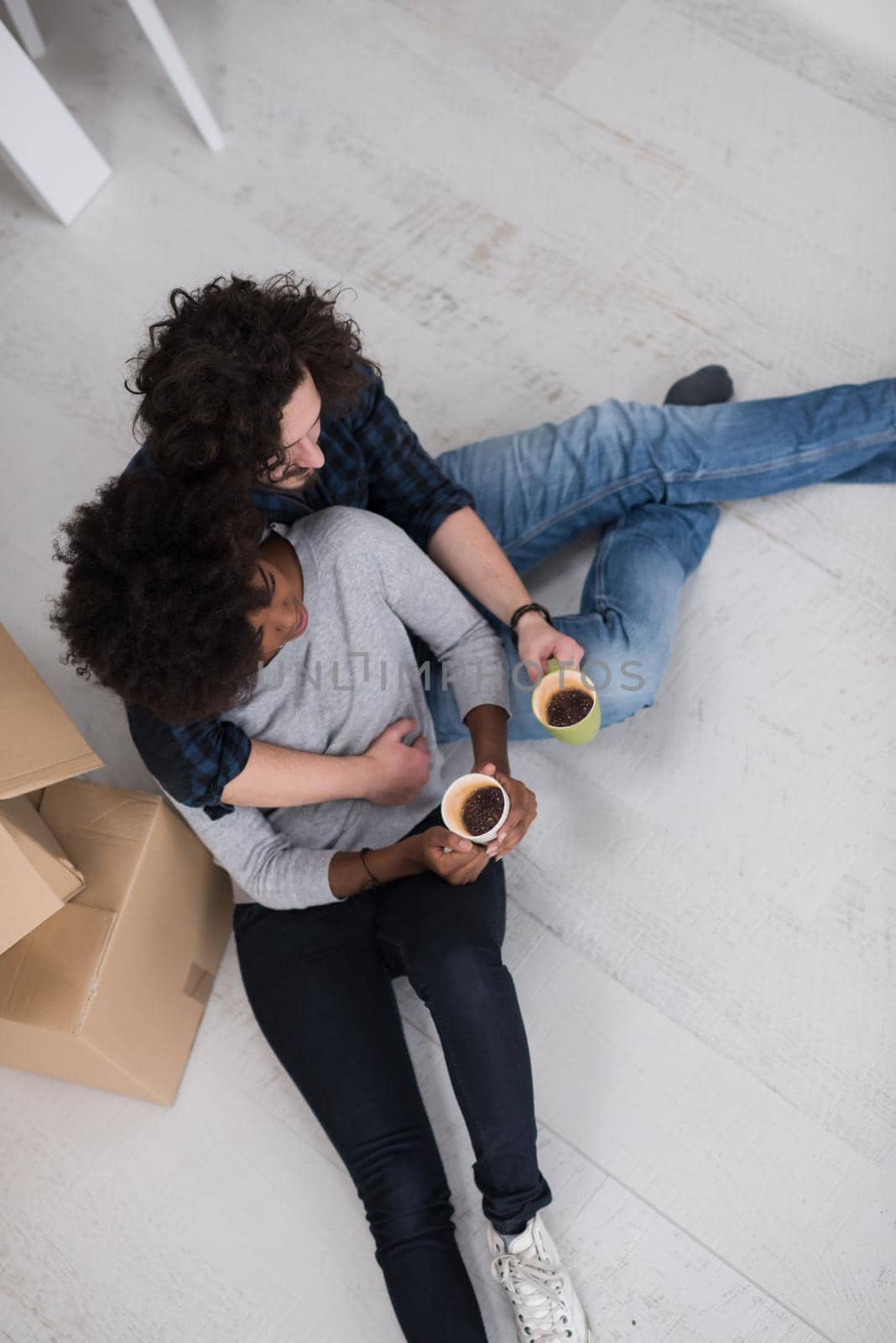 Relaxing in new house. Cheerful young multiethnic couple sitting on the floor and drinking coffee while cardboard boxes laying all around them