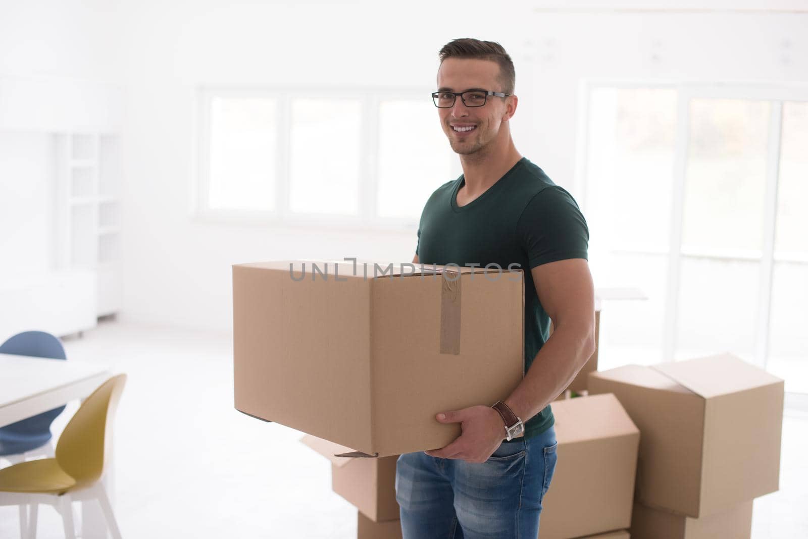 Moving to a new apartment. Cheerful young man holding a cardboard boxes and smiling at camera while other carton boxes laying on background