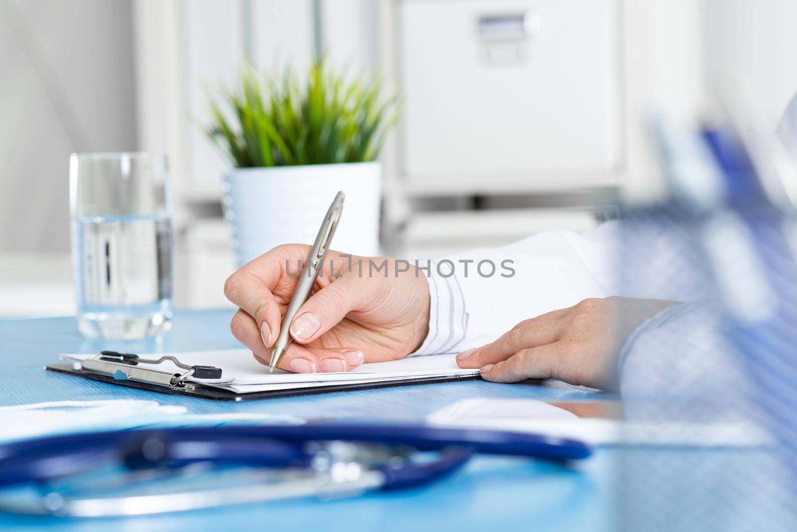 Doctor filling patient history list at clipboard. Physician in medical uniform working at desk in office. Examination and diagnosis in clinic. Doctor workplace with green plant in pot and stethoscope.