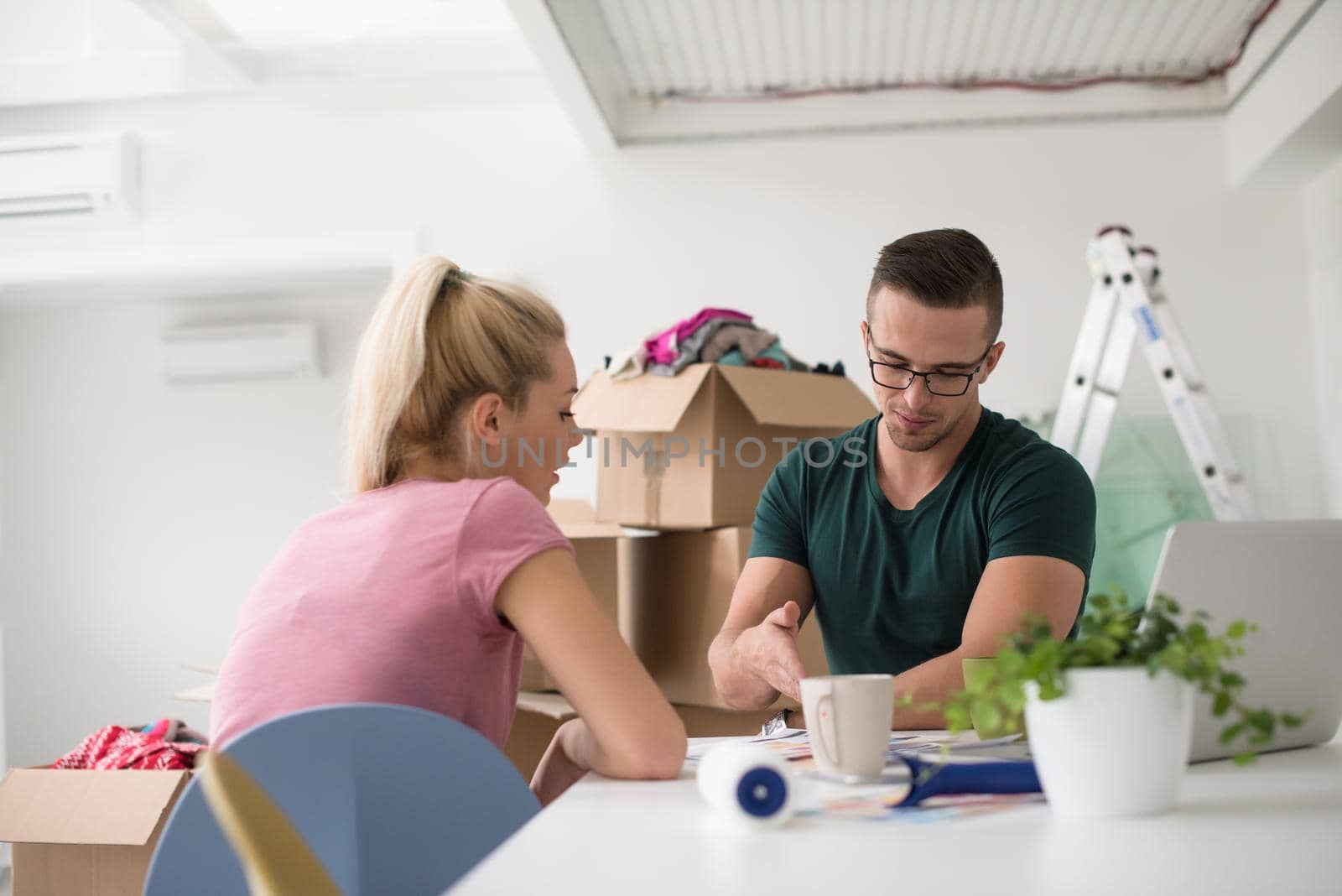 Young couple moving in a new home. Man and woman at the table using notebook laptop computer and plans with boxes around them