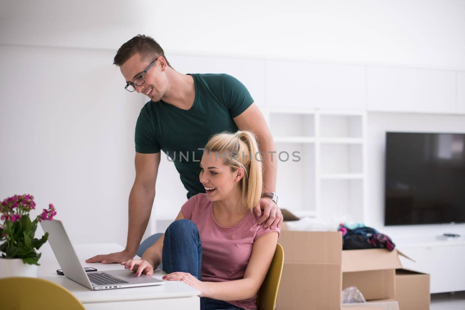 Young couple moving in a new home. Man and woman at the table using notebook laptop computer and plans with boxes around them