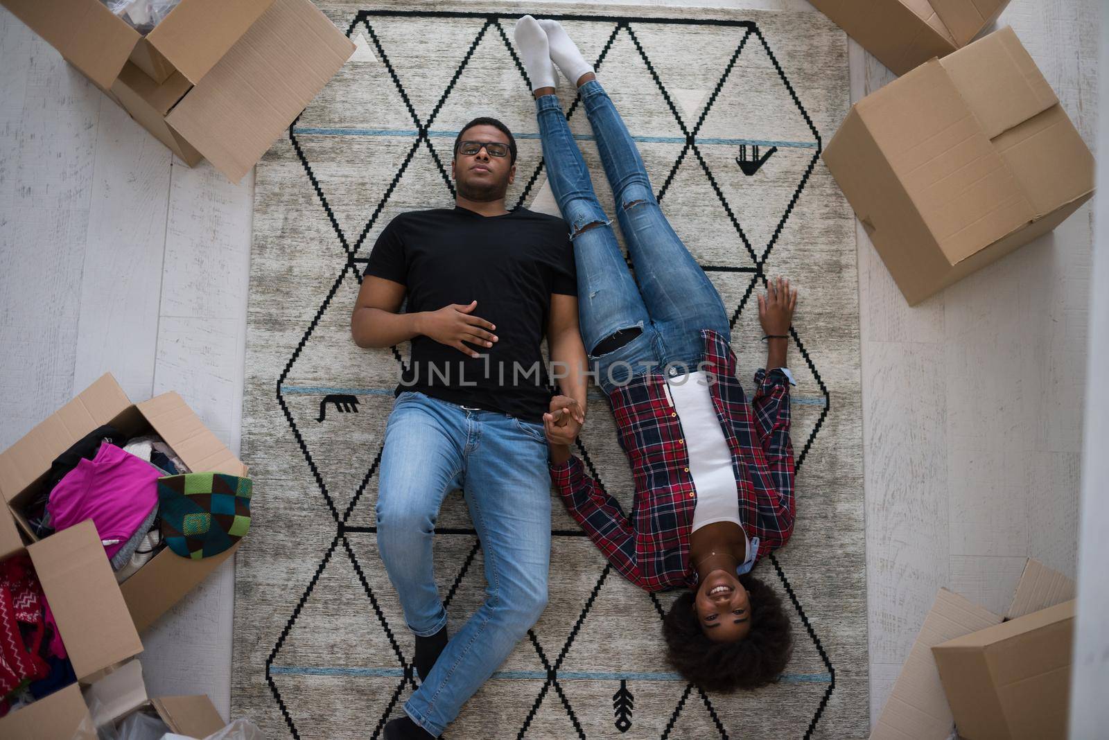 Top view of attractive young African American couple moving, holding hands, looking at camera and smiling while lying among cardboard boxes