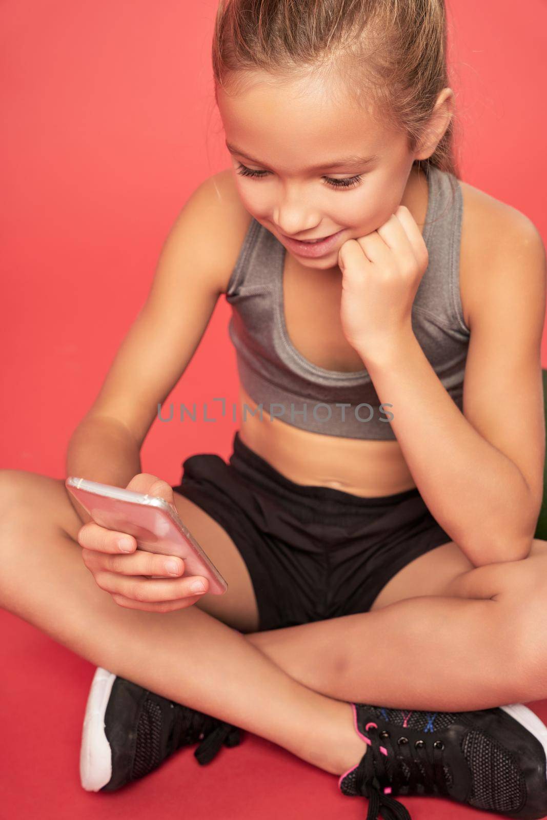 Cute female child reading message on cellphone and smiling while sitting with her legs crossed. Isolated on red background