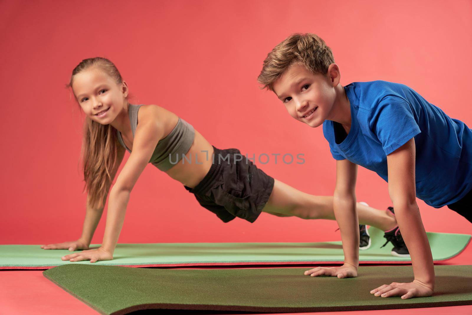 Adorable girl and boy in sportswear holding push-up position and smiling while doing strength exercise on yoga mats