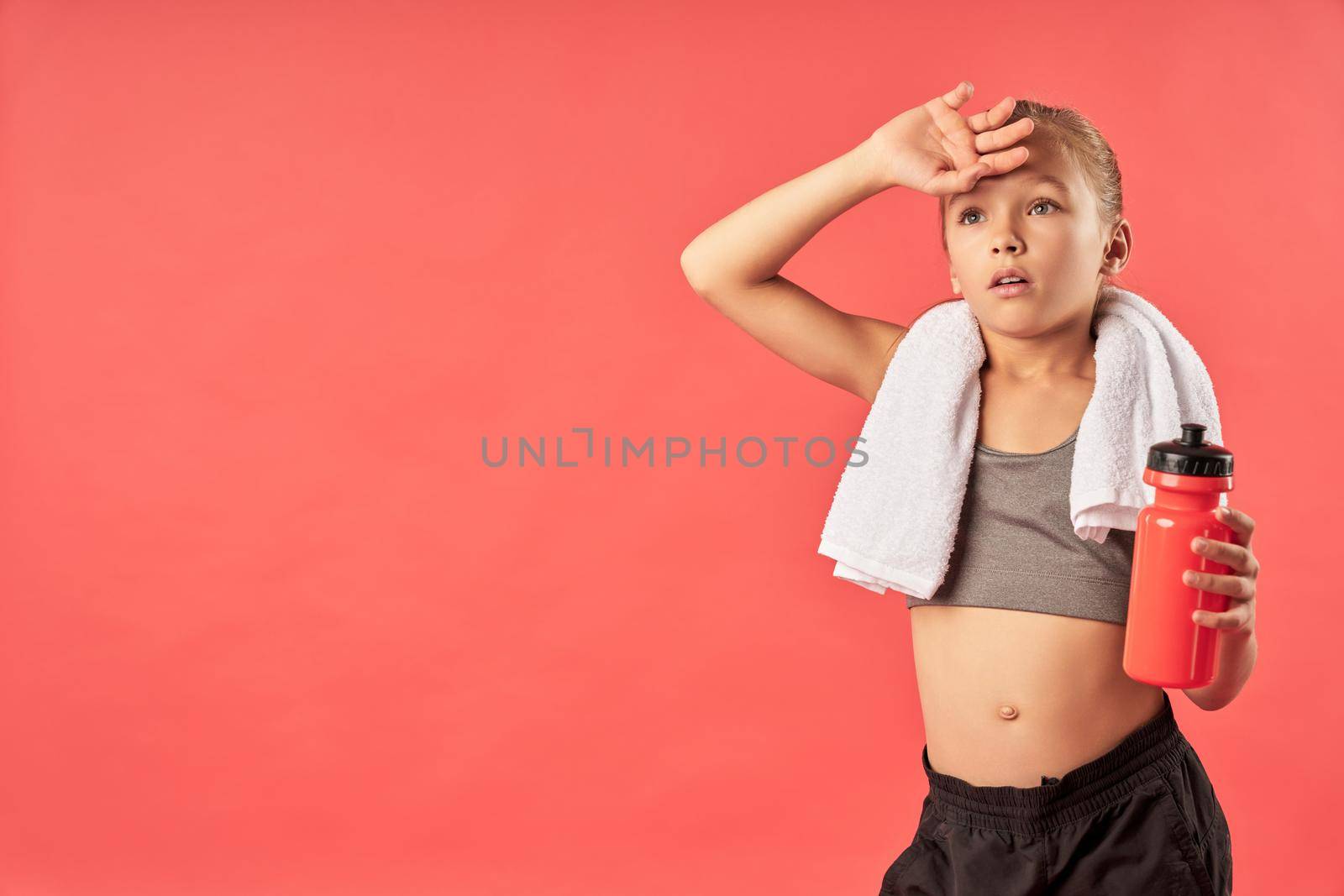 Adorable female child with towel on her shoulders holding bottle of water while standing against red background