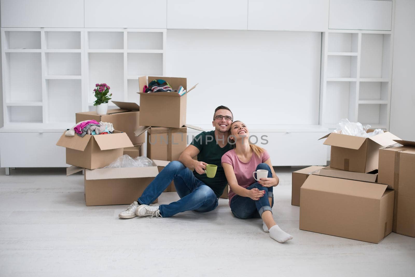 Relaxing in new house. Cheerful young couple sitting on the floor and drinking coffee while cardboard boxes laying all around them