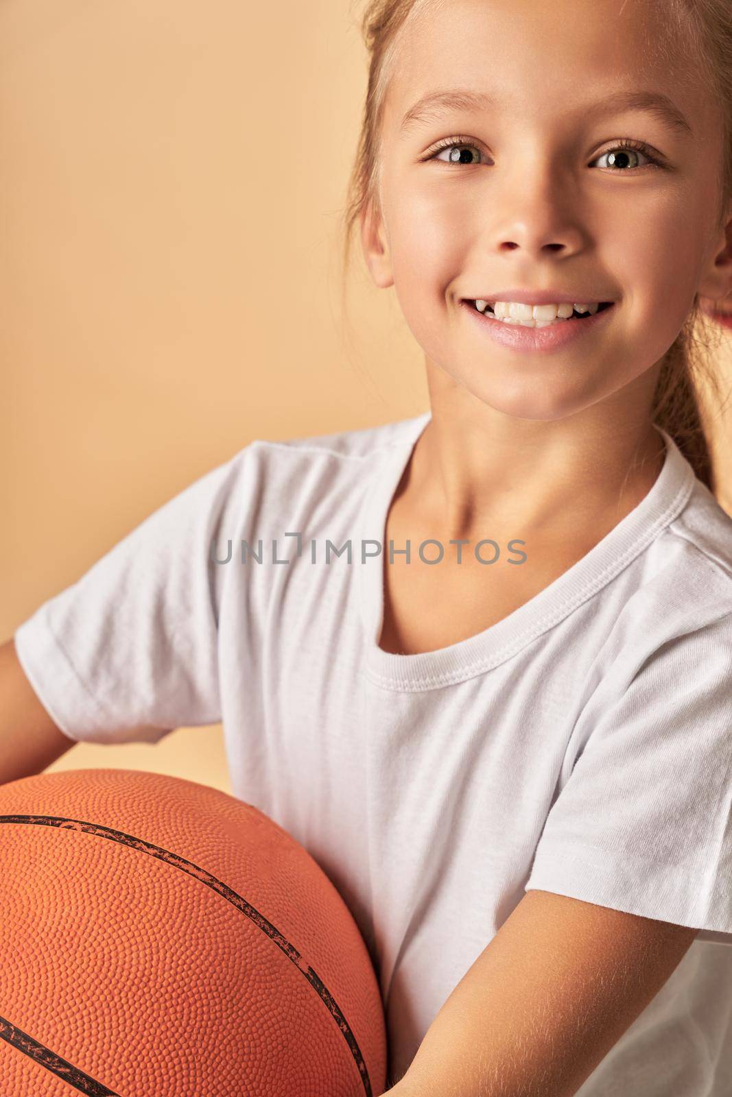 Close up of cute female child with basketball ball standing against light orange background